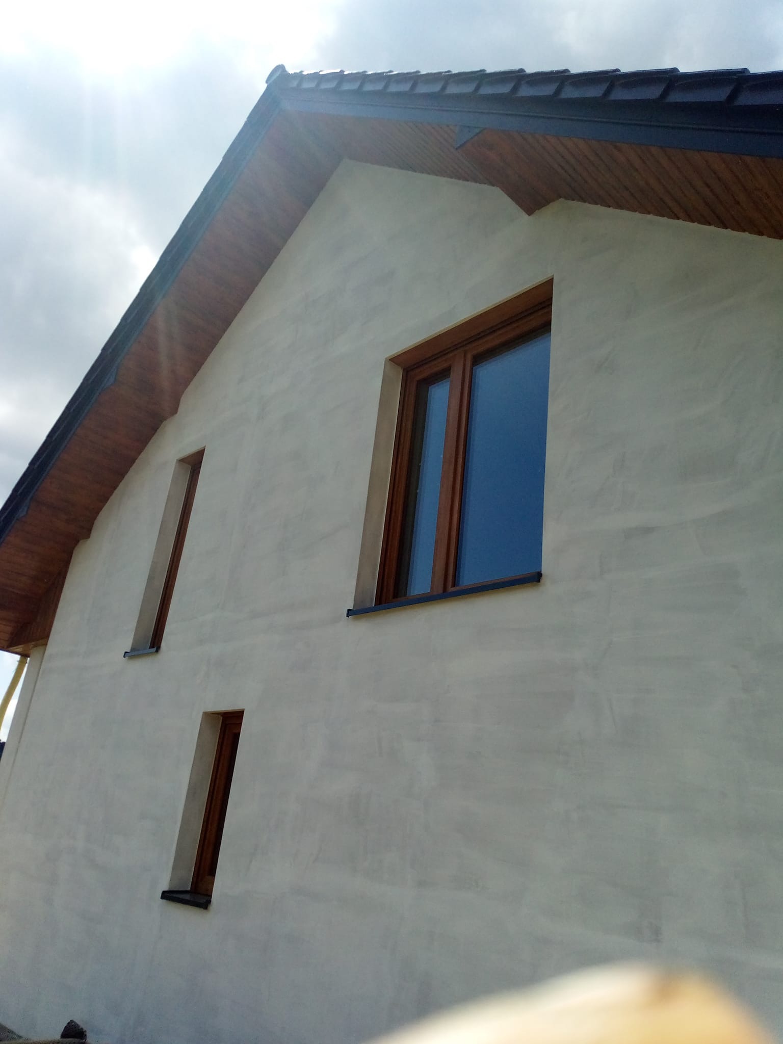 Jasna elewacja domu jednorodzinnego z drewnianą stolarką okienną i podbitką dachową, widok z dołu, w tle pochmurne niebo.