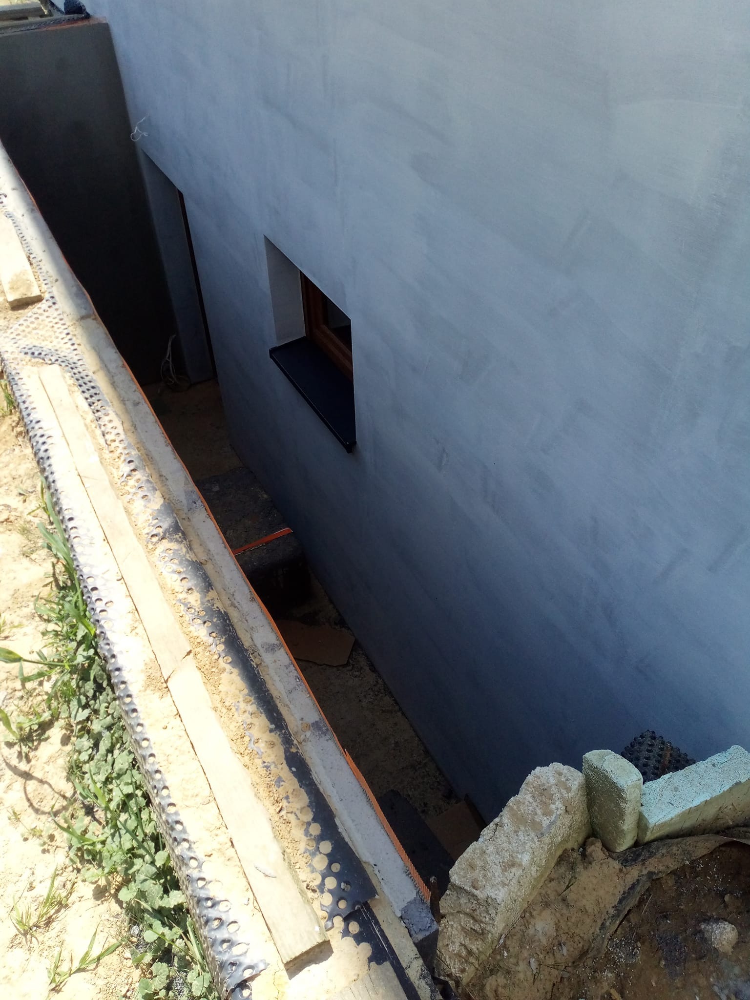 Świeżo otynkowana, szara elewacja budynku z małym oknem, widoczna z poziomu gruntu. W tle ciemne wejście. Na pierwszym planie elementy konstrukcyjne.