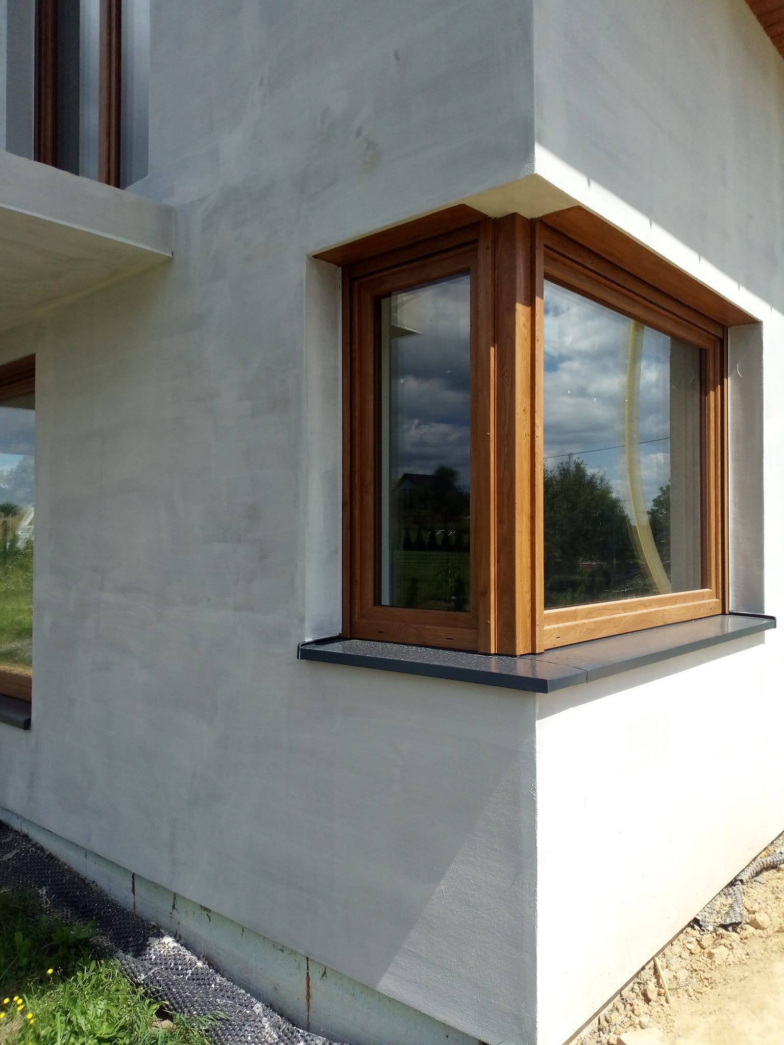 Szara elewacja domu jednorodzinnego z drewnianą stolarką okienną i czarnym parapetem, widok z zewnątrz, fragment budynku z oknem narożnym.