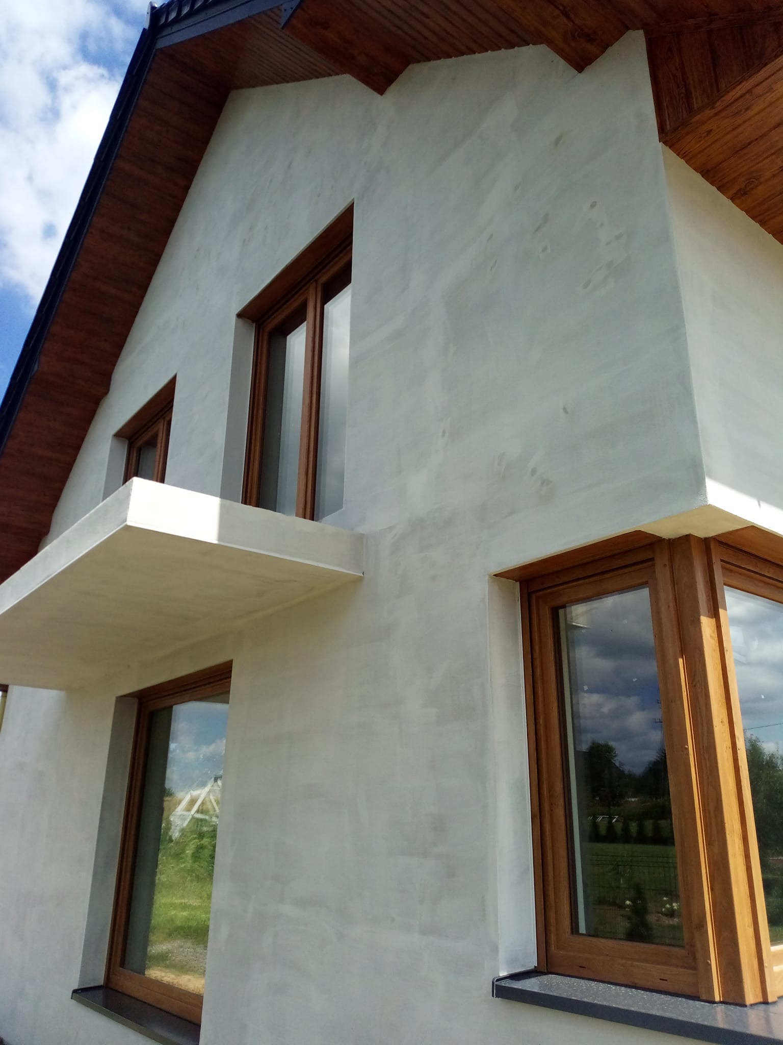 Nowoczesny dom z jasną elewacją o fakturze baranka i drewnianymi oknami. Widok z dołu, podkreślający bryłę budynku i detale wykończenia.