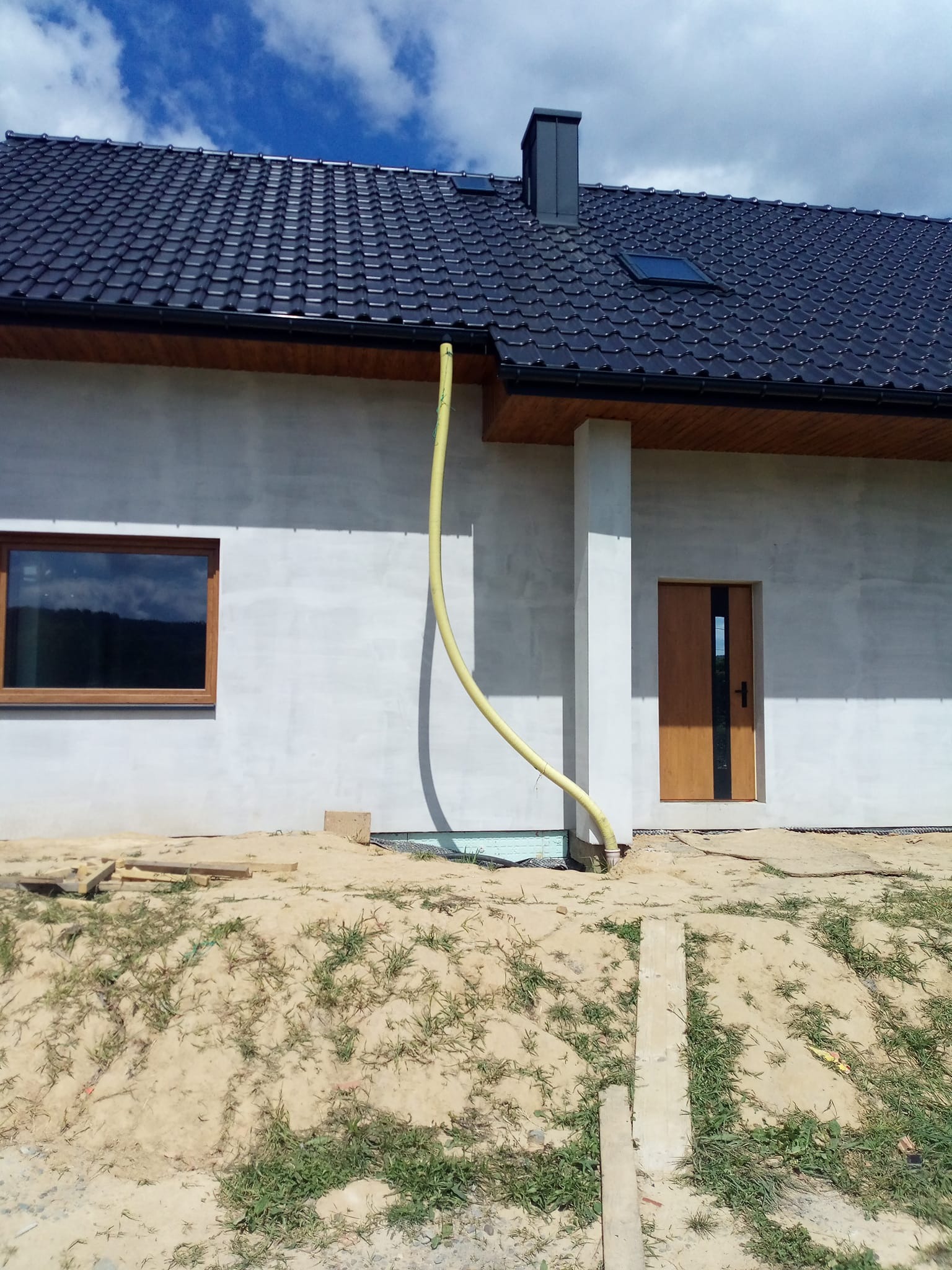 Surowa elewacja domu jednorodzinnego z ciemną dachówką, widoczne okno, drzwi i żółta rura spustowa. Teren wokół domu w trakcie prac ziemnych.