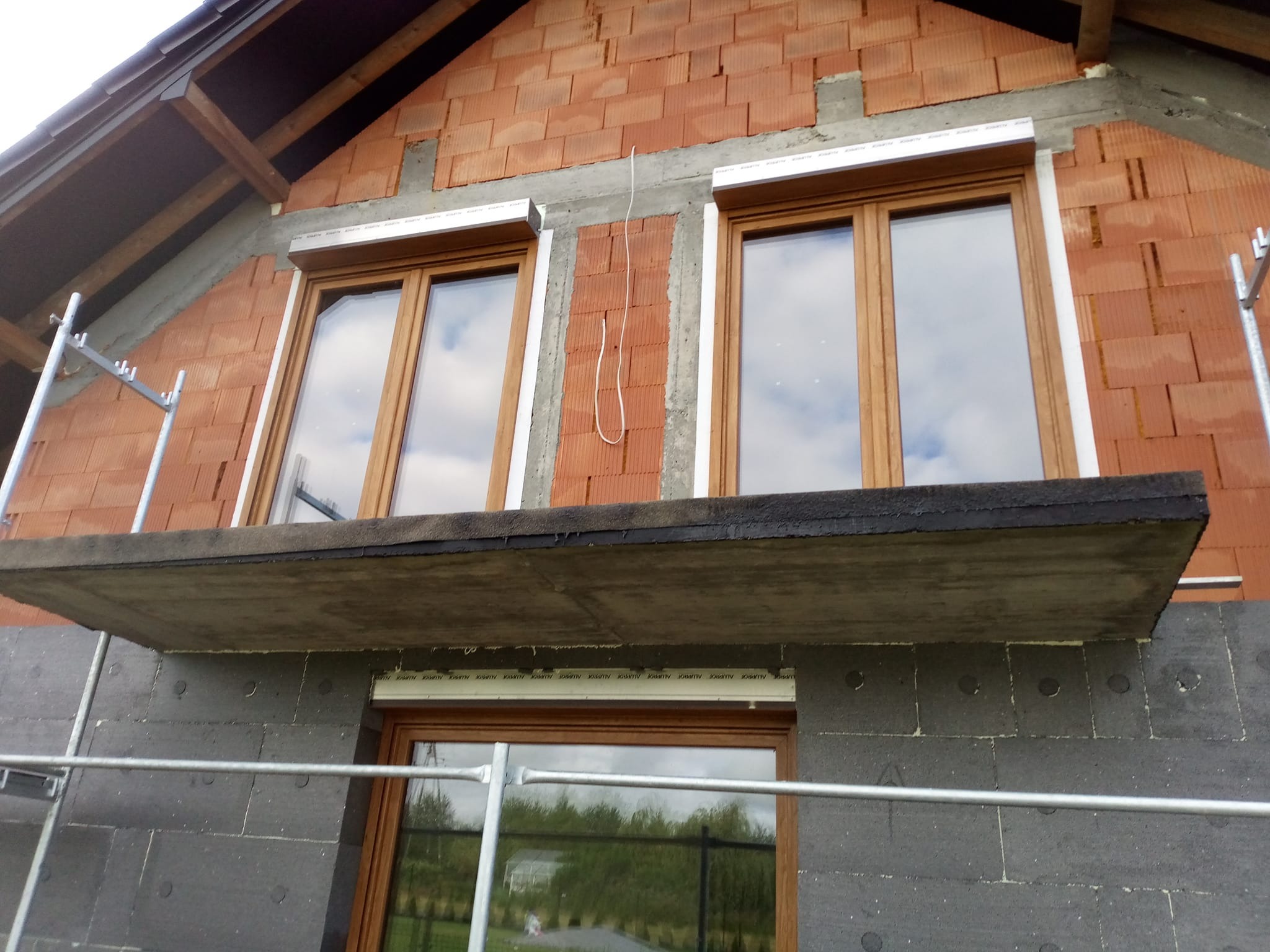 Budynek w trakcie prac elewacyjnych: widoczne okna z roletami, fragment rusztowania i ocieplenie z szarego styropianu. Część ściany z czerwonej cegły.