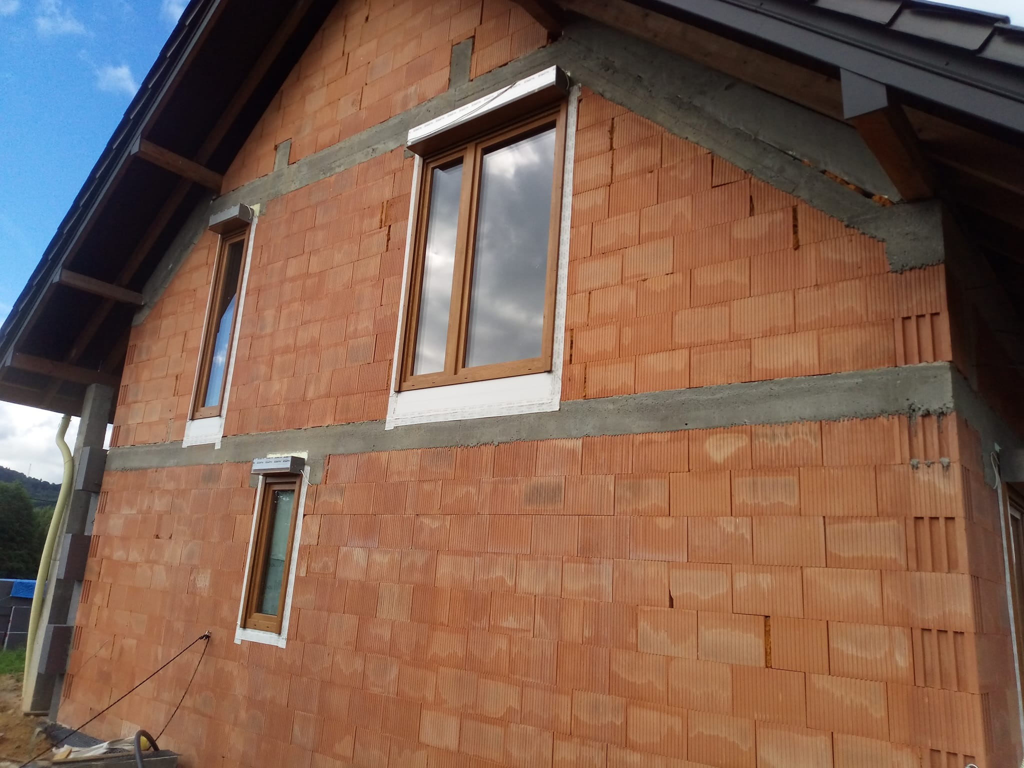 Surowa elewacja z czerwonej cegły ceramicznej z zamontowanymi oknami z brązowymi ramami. Widoczne betonowe nadproża i fragment dachu. Budynek w trakcie budowy.