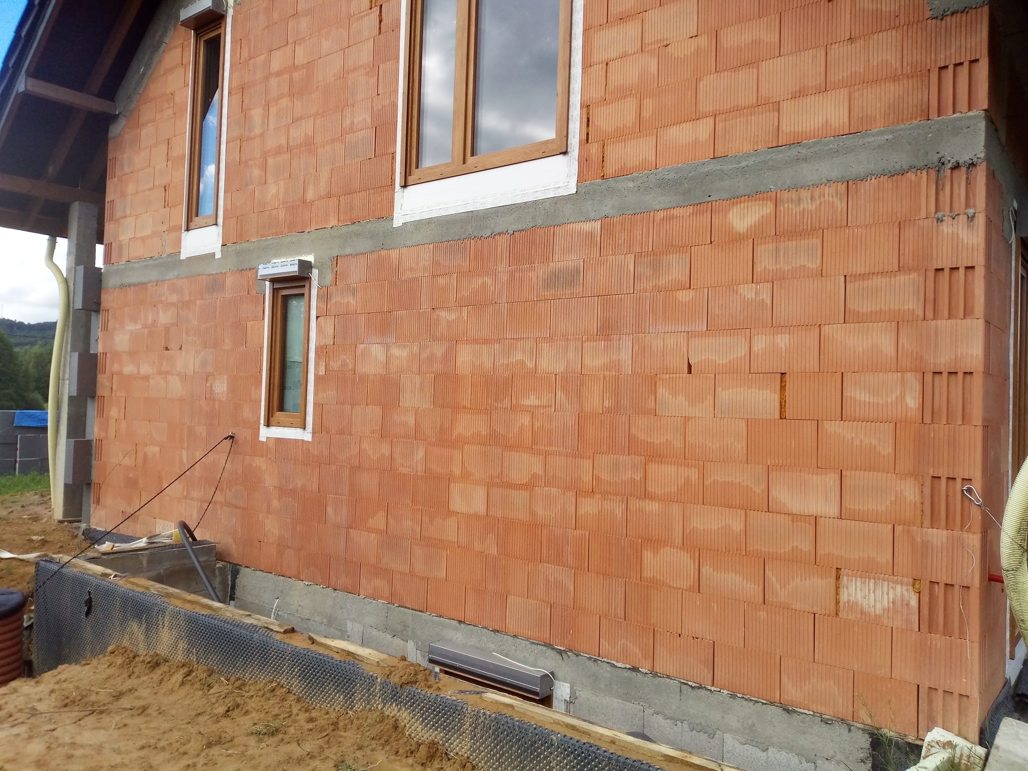 Ściana domu w trakcie budowy z czerwonej cegły, widoczne okna z drewnianymi ramami i fundamenty. Etap murowania ścian.