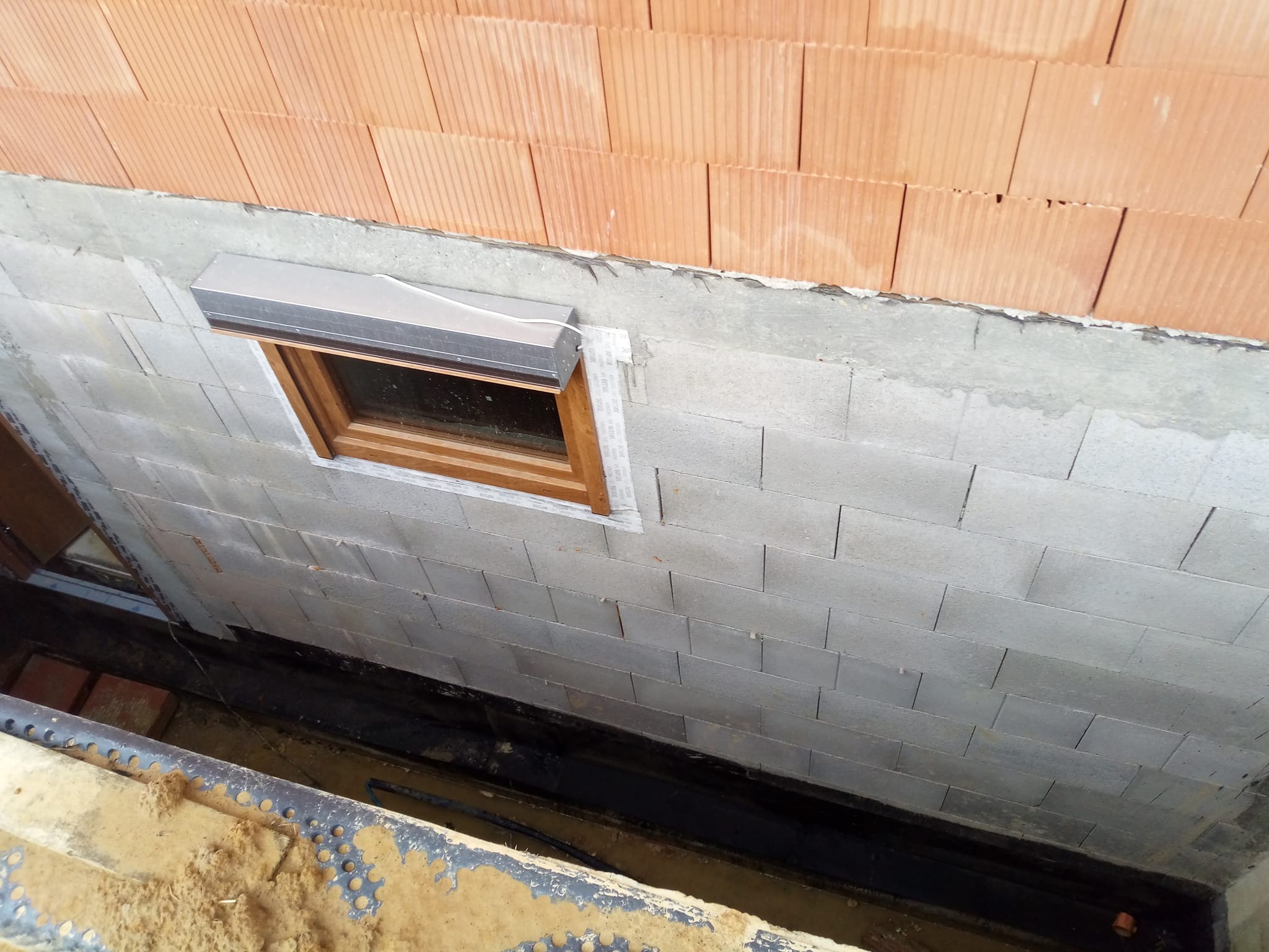 Ściana z szarego bloczka z oknem i fragmentem czerwonej cegły powyżej. Widoczna czarna izolacja i fragment wykopu z piaskiem. Etap budowy elewacji.