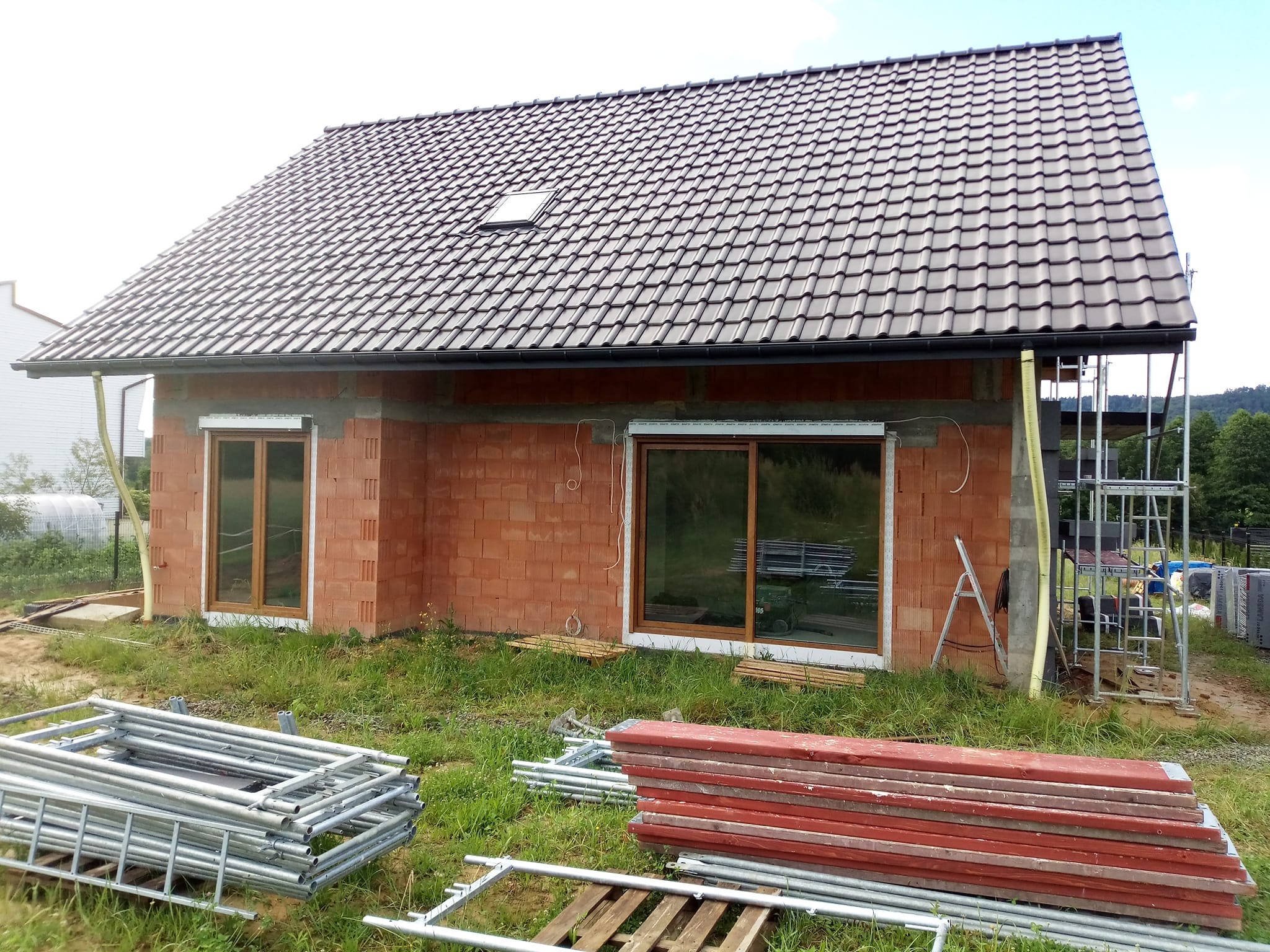 Surowy budynek z czerwonej cegły z oknami i dachem z dachówki, otoczony rusztowaniami i materiałami budowlanymi na trawie. Widoczne elementy instalacji.