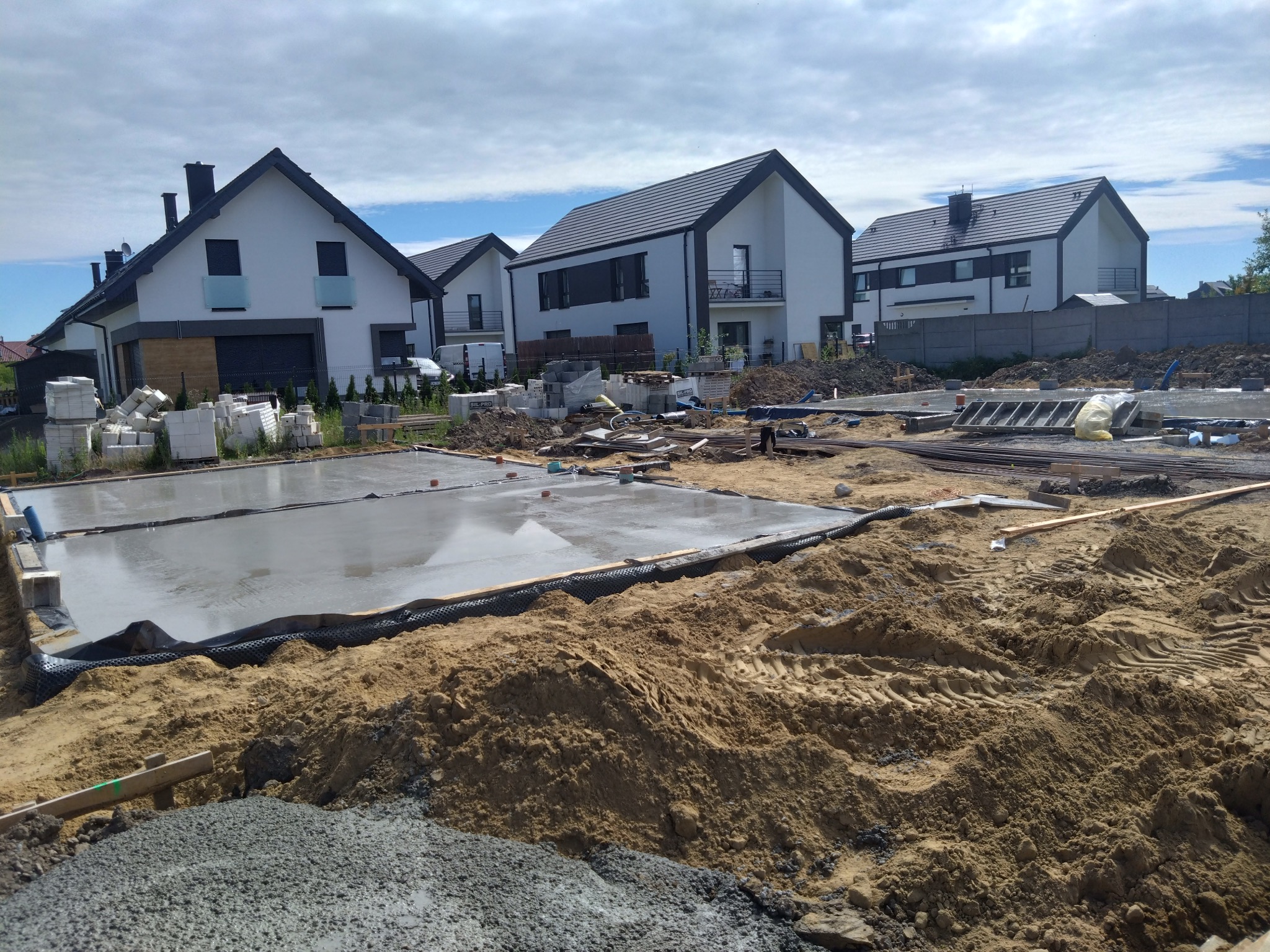 Świeżo wylana, gładka wylewka fundamentowa na budowie osiedla domów jednorodzinnych w nowoczesnym stylu, z widocznym zbrojeniem i deskowaniem.