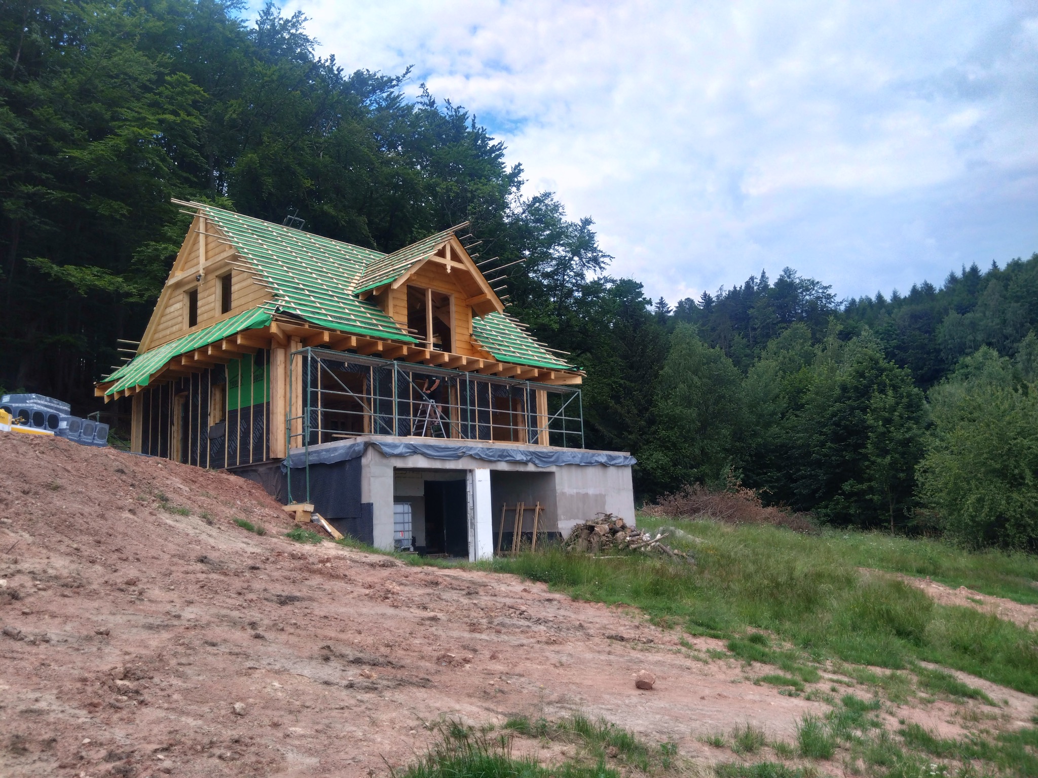 Drewniany dom w trakcie budowy na wzgórzu, z zielonym dachem i widocznym szkieletem konstrukcji. W tle las i pochmurne niebo. Widoczny betonowy fundament.