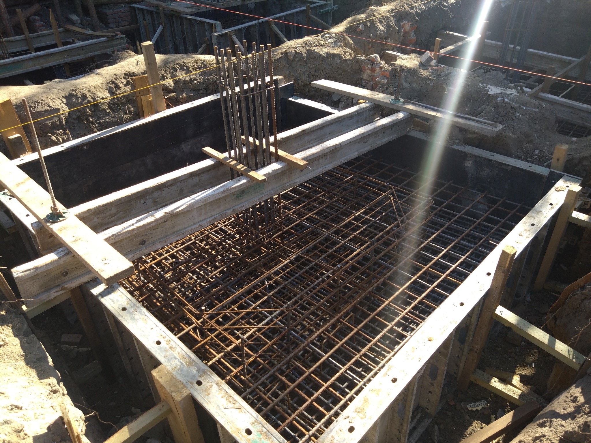 Zbrojenie fundamentu w szalunku z desek, widok z góry. Stalowe pręty tworzą siatkę, wystające pionowe elementy. Świeże wykopy ziemne w tle.