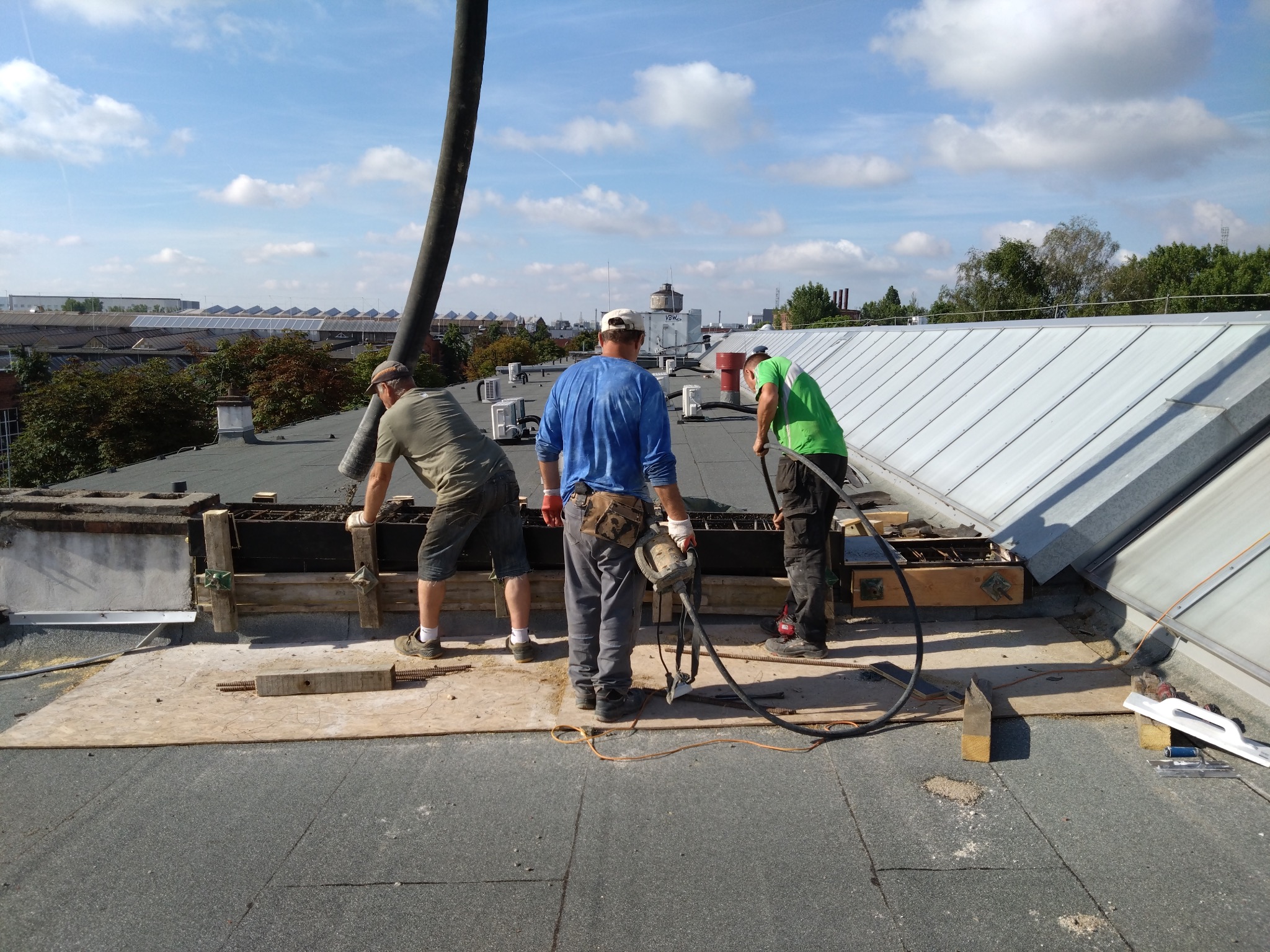 Trzech robotników na dachu budynku podczas prac remontowych. Widoczna konstrukcja szalunkowa, narzędzia i elementy zbrojenia. W tle zabudowa miejska i błękitne niebo.