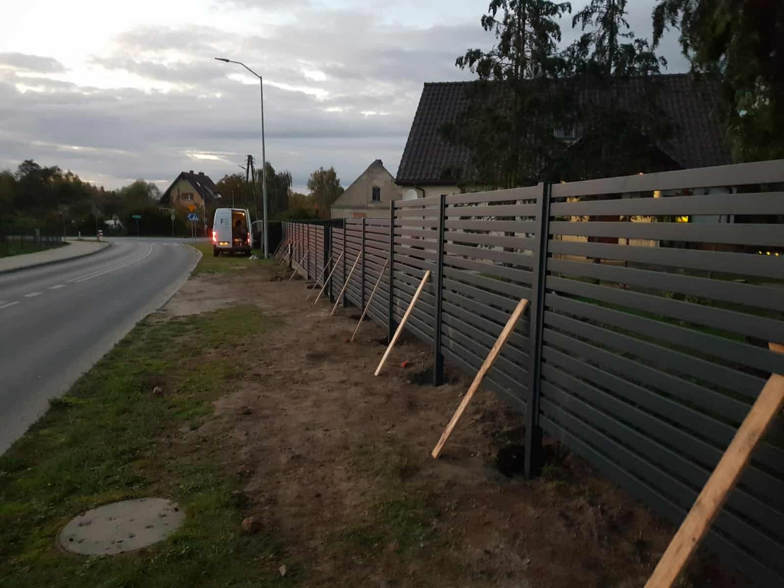 Nowoczesne ogrodzenie panelowe w Wałczu, w trakcie montażu. Widoczne podpory stabilizujące, szara kolorystyka paneli i słupków. W tle droga i zabudowa mieszkalna.