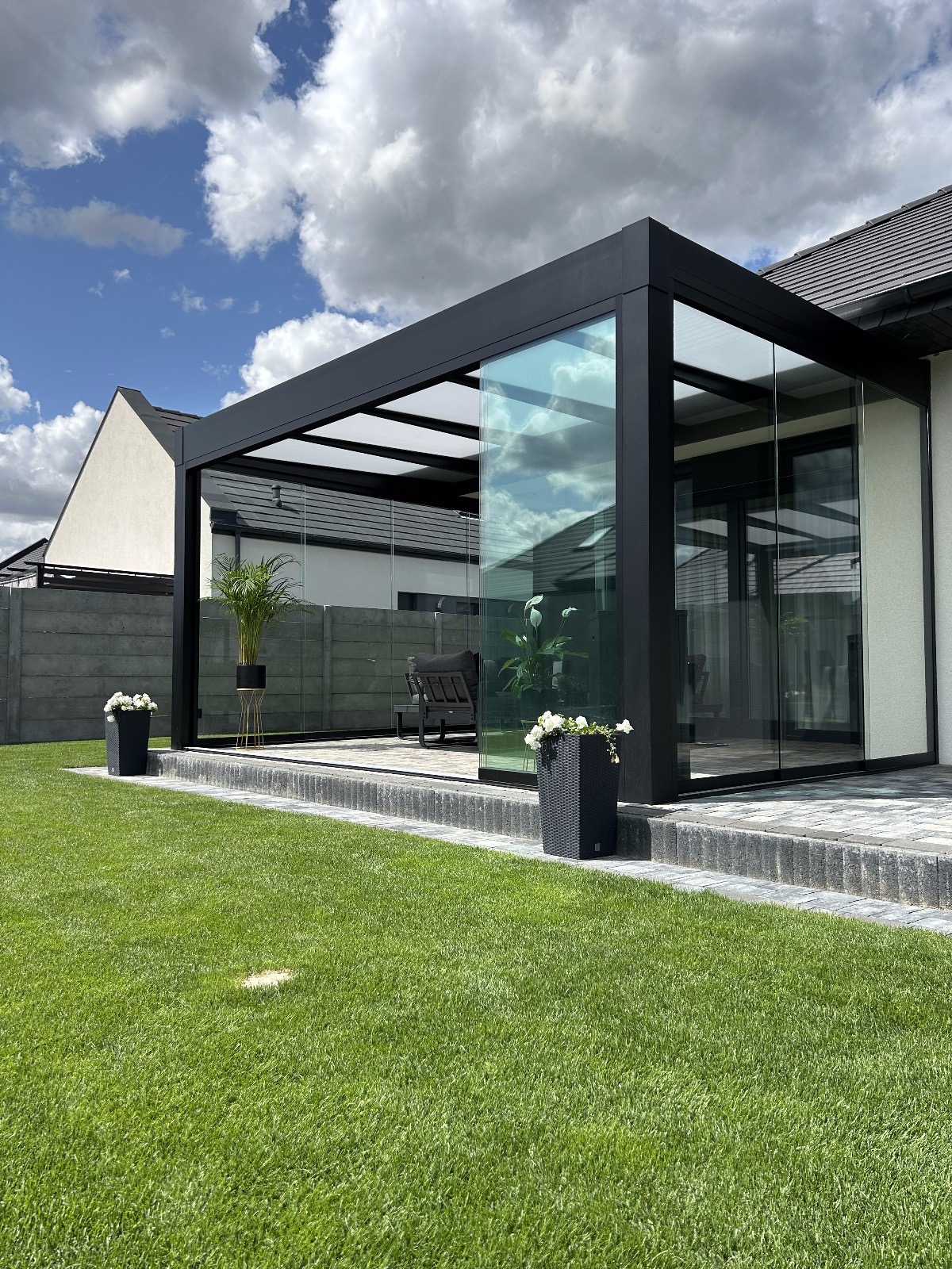 Nowoczesna, przeszklona pergola przyścienna z czarną ramą i mlecznym poliwęglanem na dachu, otoczona zielonym trawnikiem i dekoracyjnymi donicami.