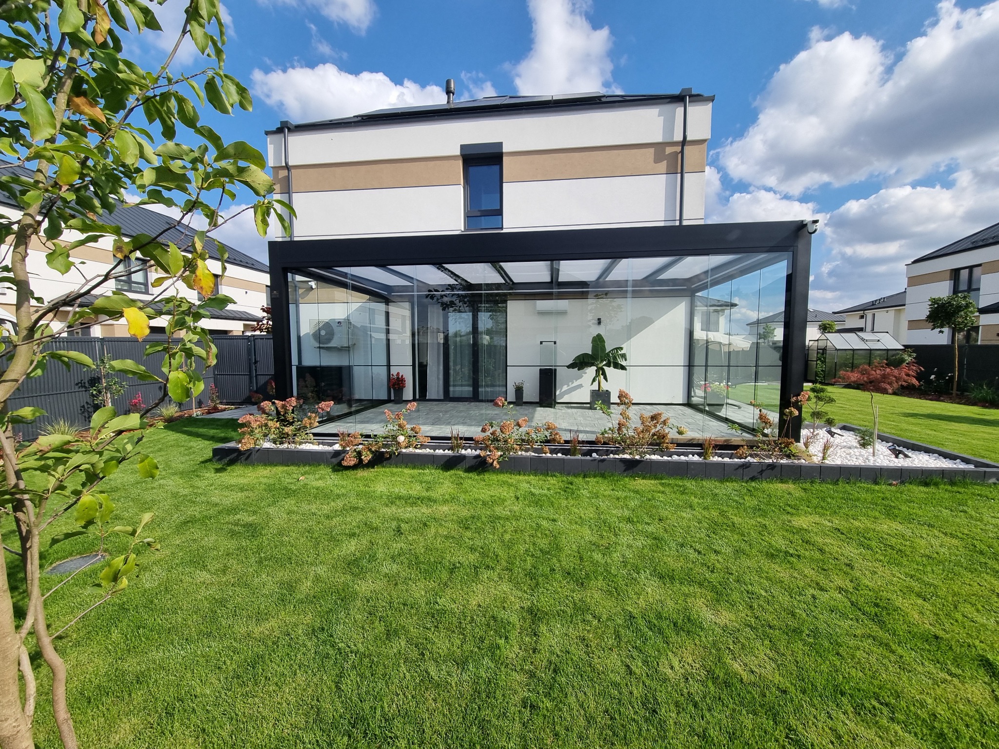 Szklana zabudowa tarasu przy domu w Golina. Zadaszenie przyścienne z poliwęglanowym dachem i szklanymi ścianami. Widok na zadbany trawnik i rośliny.