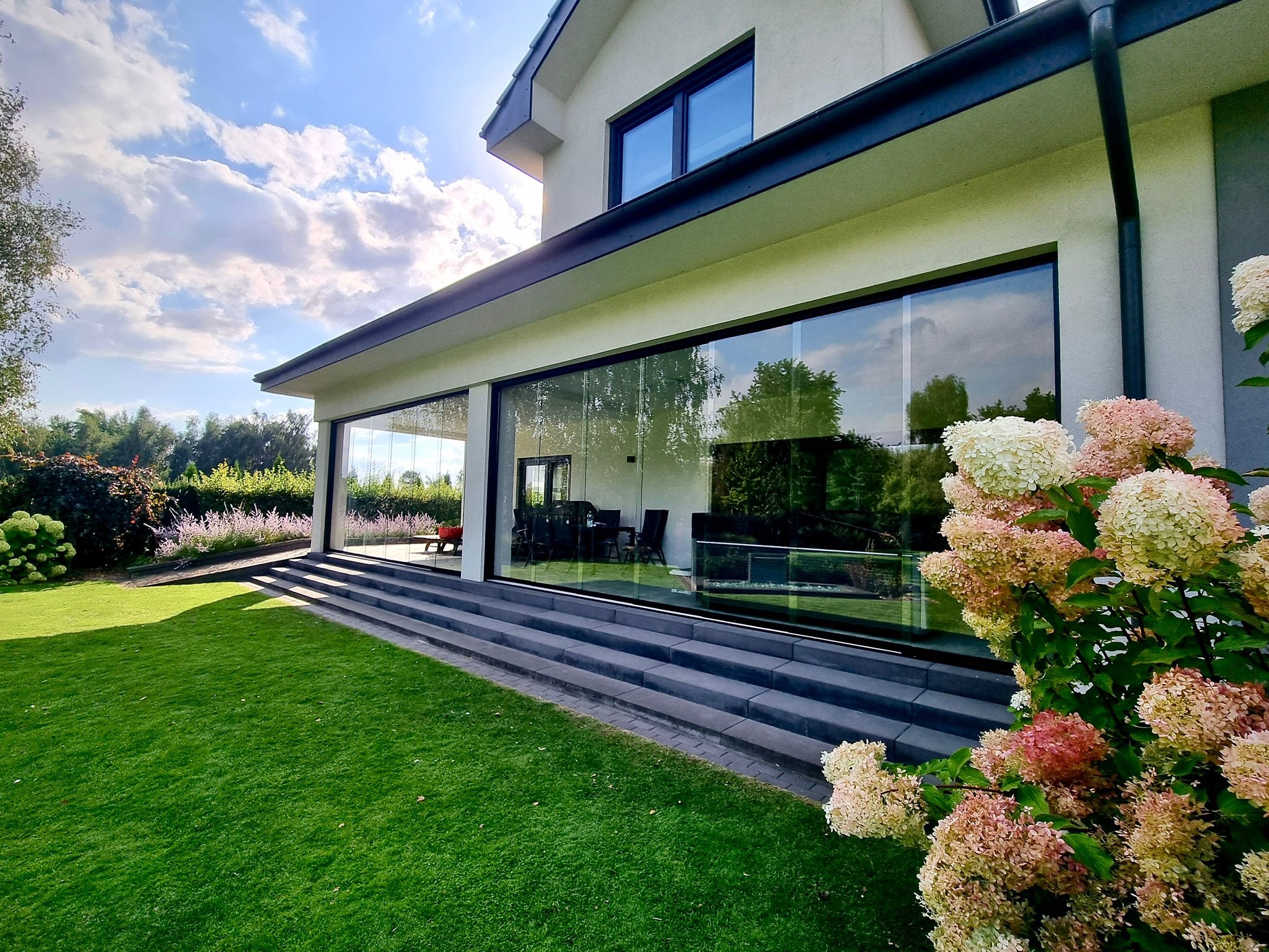 Nowoczesny taras z przesuwnymi szklanymi ścianami, widok z zewnątrz na zadbany trawnik i hortensje. Słoneczny dzień.