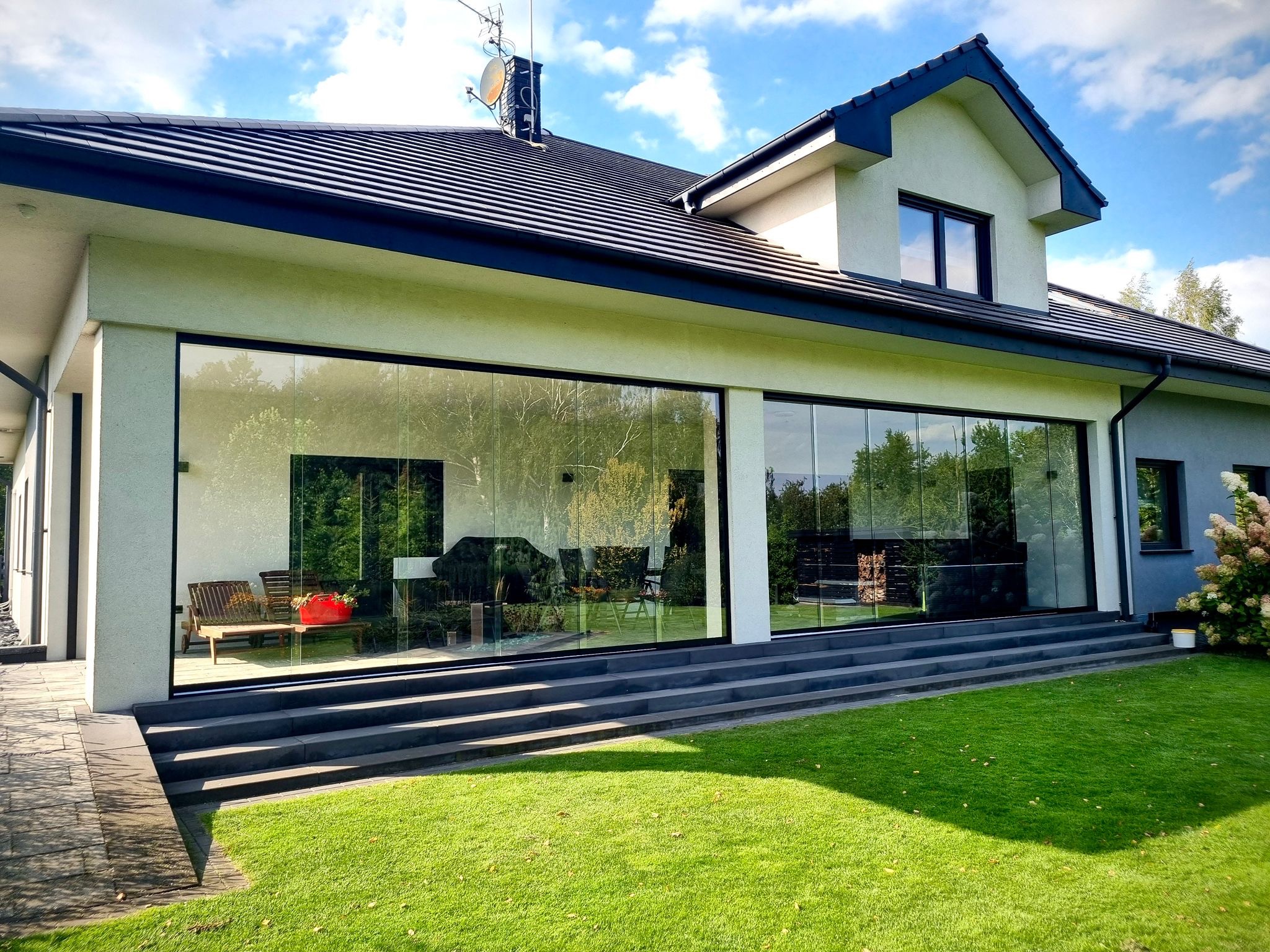 Nowoczesny dom z przeszklonym tarasem i przesuwnymi szybami, widok z zewnątrz na zadbany trawnik i schody.