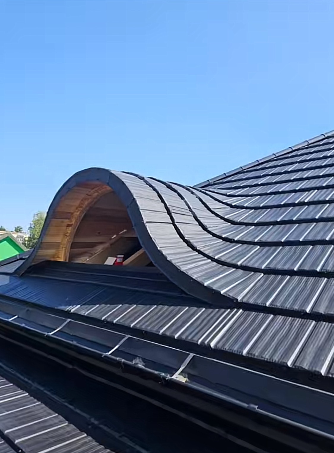 Falista blacha na dachu z drewnianym lukarną w kształcie łuku. Nowoczesne pokrycie dachowe w odcieniach szarości, widok z dołu na tle błękitnego nieba.