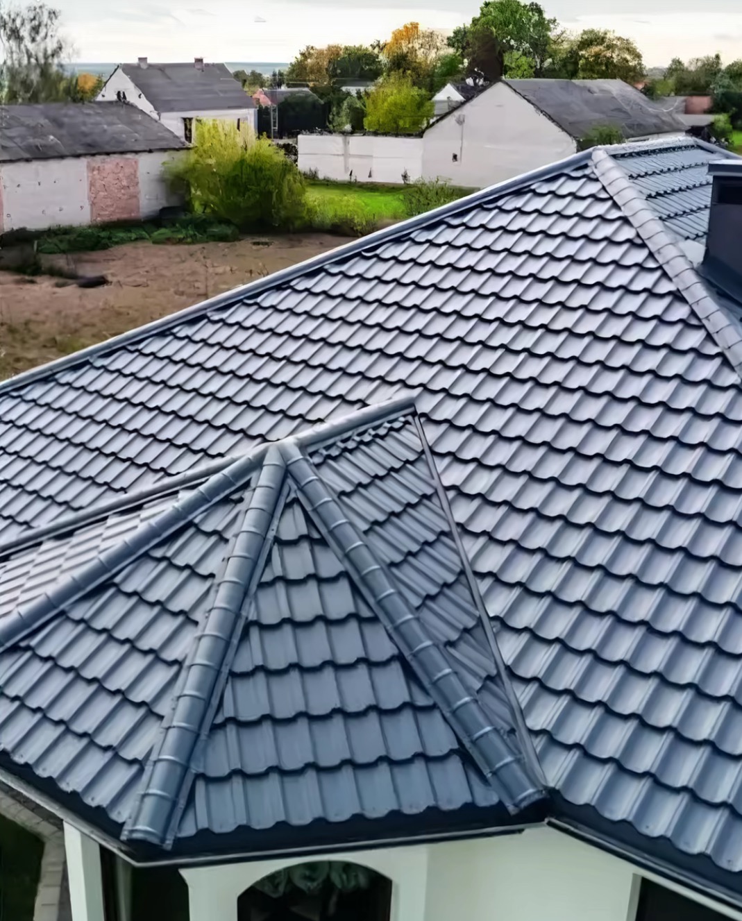 Szary dach z blachy na domu jednorodzinnym, widok z góry. Charakterystyczne ułożenie blachy tworzy wzór. W tle zabudowa mieszkalna i zieleń.