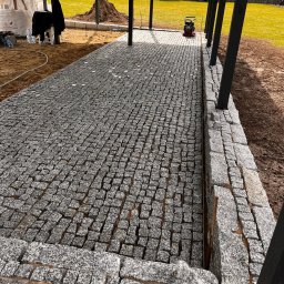 Art_Stone - Świeżo ułożona, szara kostka granitowa pod zadaszeniem, z widoczną ubijarką i przygotowanym terenem. Widok na fragment ogrodu i ogrodzenie w tle.