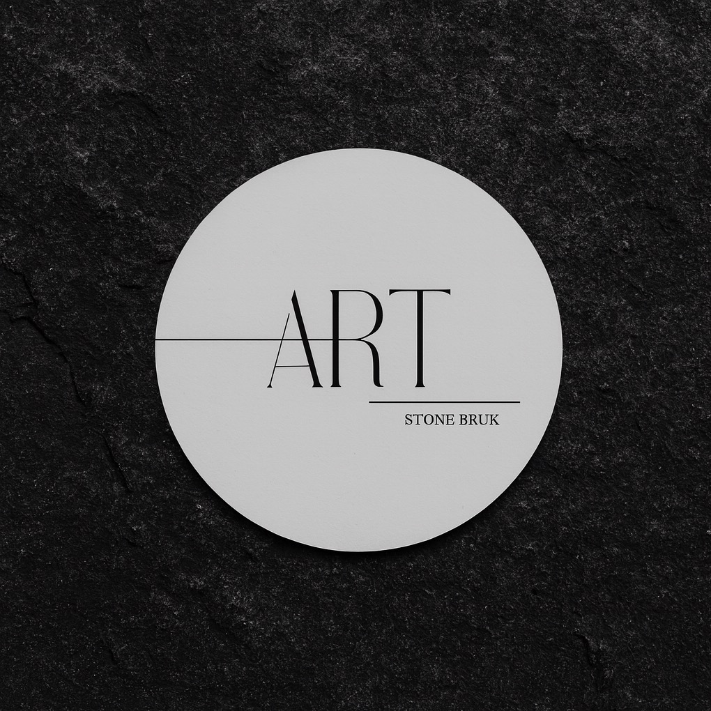 Eleganckie logo 'ART STONE BRUK' na okrągłej, jasnej etykiecie, umieszczone na tle z ciemnego, naturalnego kamienia o wyraźnej fakturze.