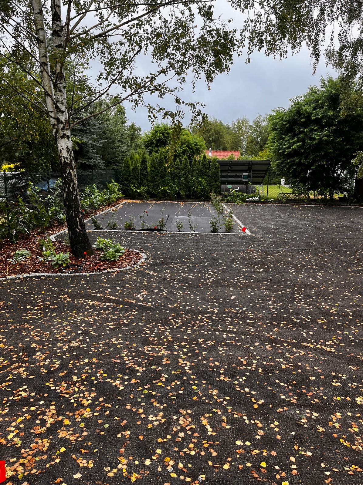 Ułożona, czarna nawierzchnia z kostki brukowej, obsypana liśćmi, z obrzeżem z kostki granitowej i nasadzeniami. W tle drzewa i budynek.