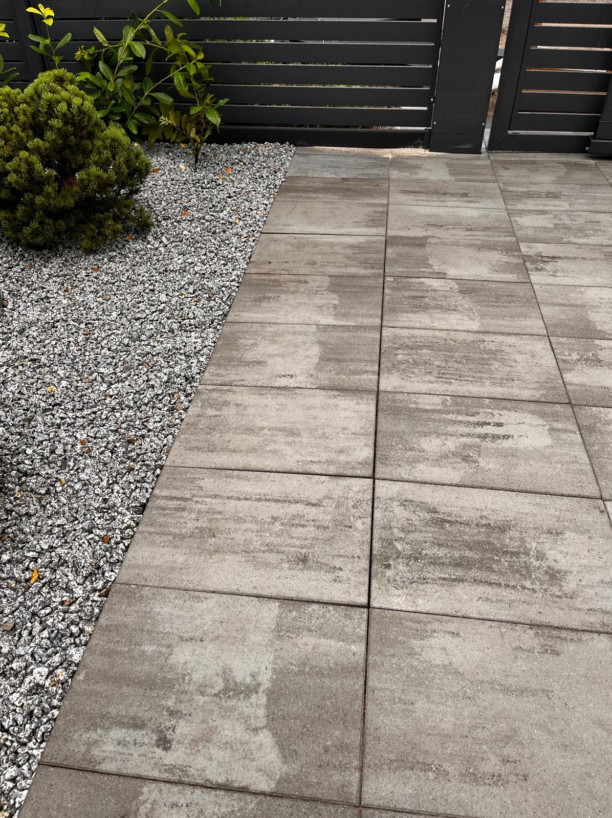 Ułożona, szara kostka brukowa z delikatnym wzorem, sąsiadująca z rabatą wysypaną drobnym, szarym żwirem i niską zielenią, całość zamknięta ciemnym ogrodzeniem.