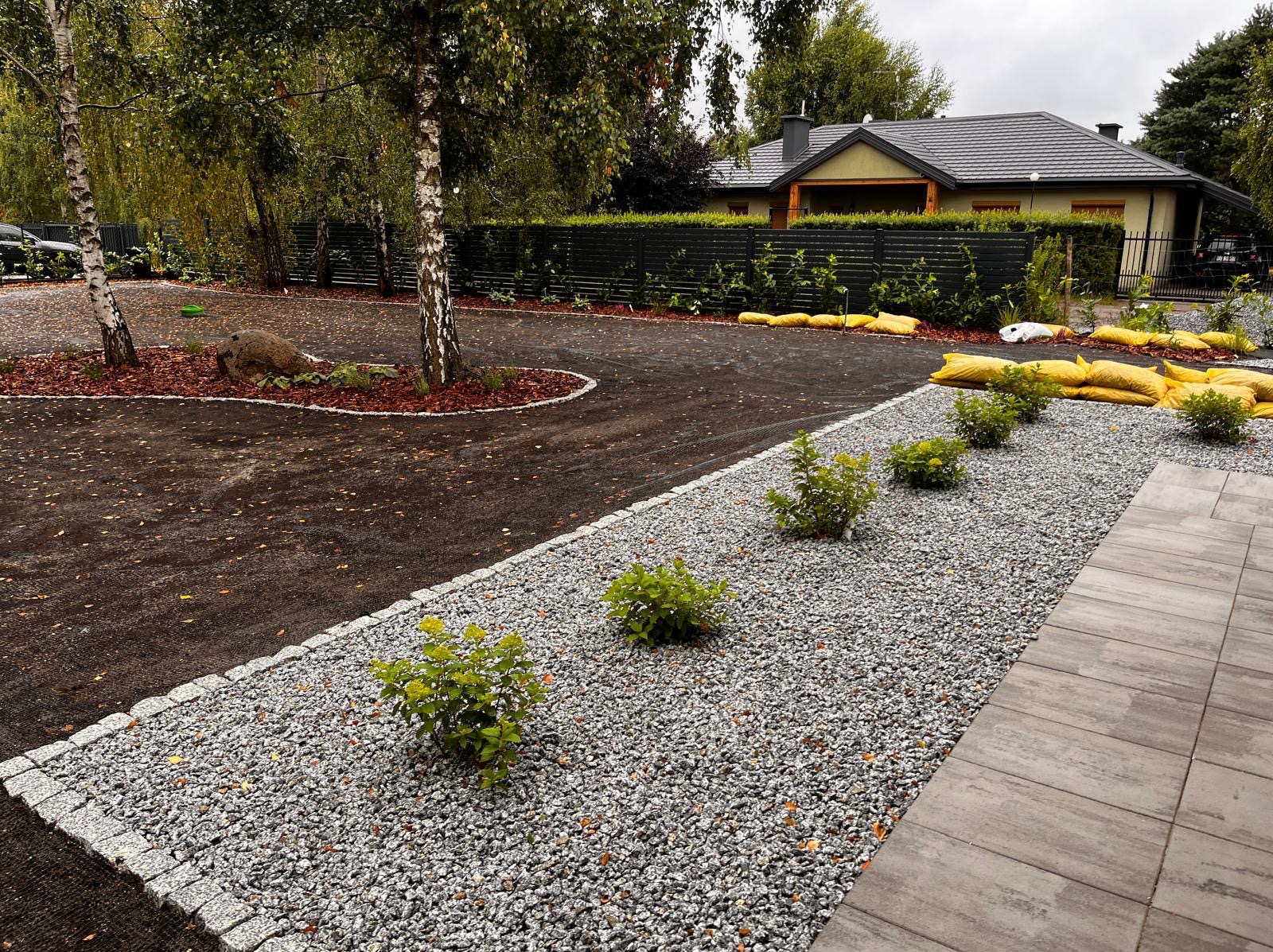 Wykończony podjazd z obrzeżami z kostki, wysypany grysem, z nasadzeniami krzewów. W tle dom z ciemną elewacją i ogrodzenie z drewnianych paneli.