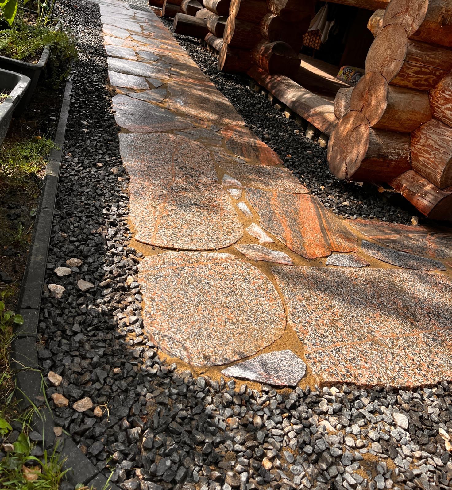 Ścieżka z nieregularnych płyt kamiennych, otoczona drobnym żwirem, prowadząca wzdłuż drewnianego domu z bali. Widoczne elementy drenażu liniowego.