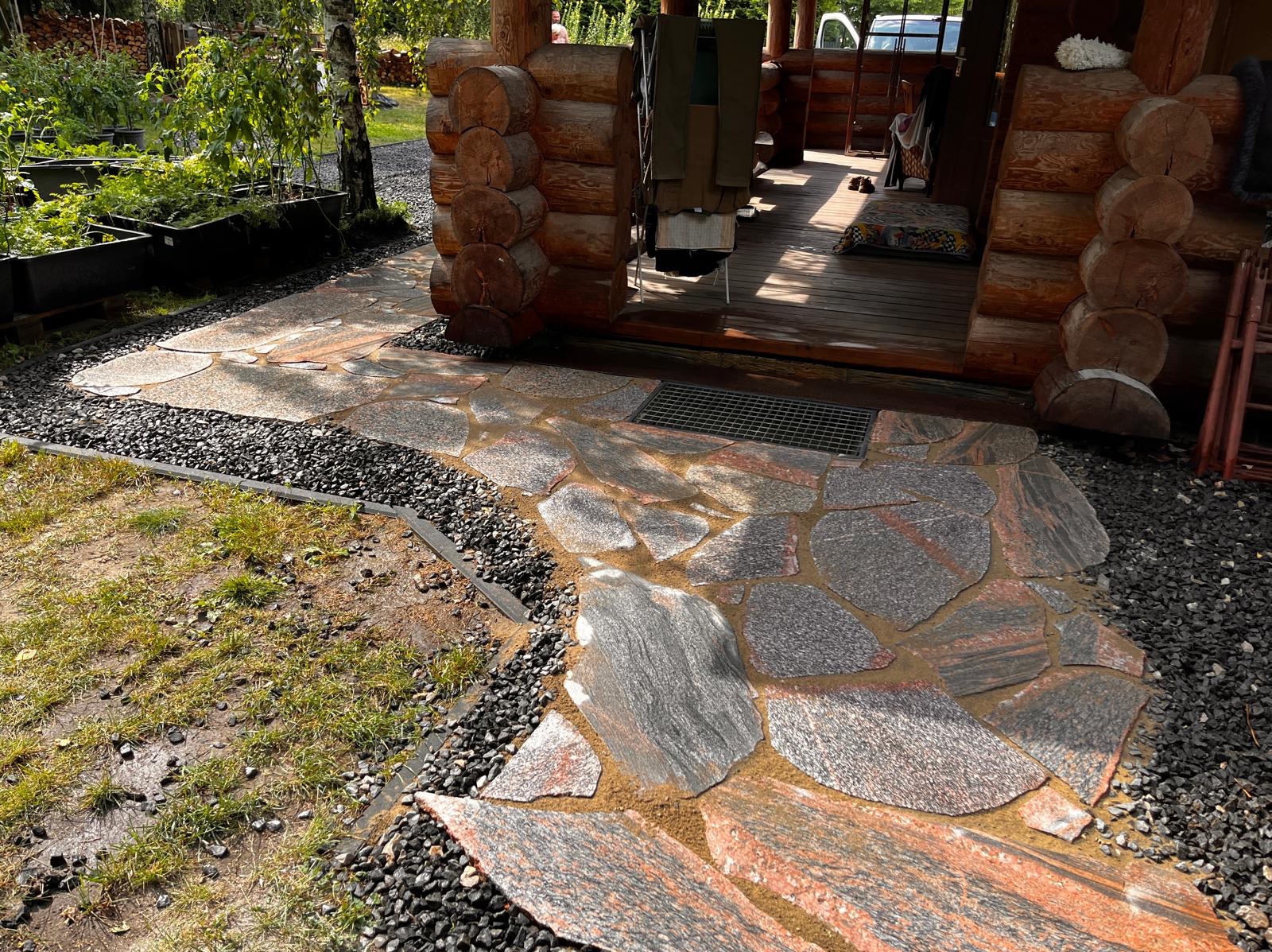 Ścieżka z nieregularnych płyt kamiennych w ogrodzie, obrzeża z czarnego grysu, drewniany budynek w tle. Widok na wejście do domu.