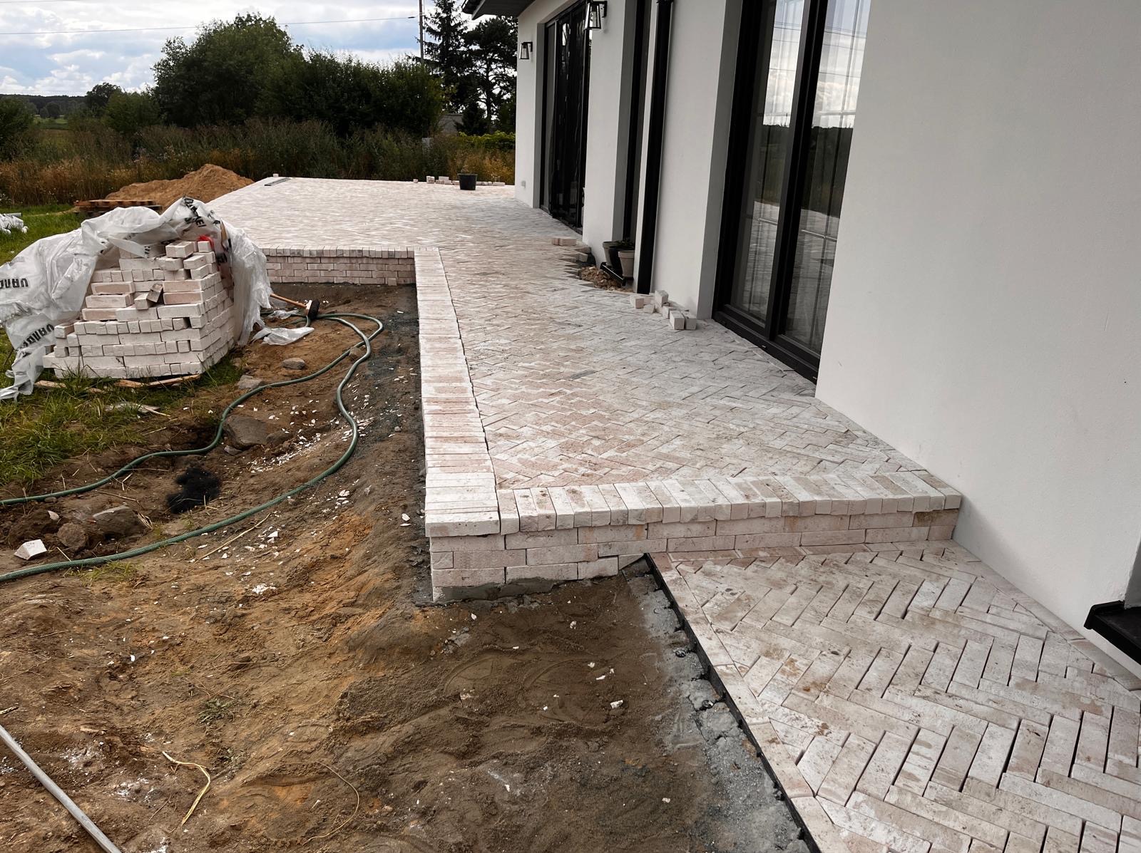 Układanie kostki klinkierowej w jodełkę na tarasie domu. Widoczne zapasy kostki, obrzeża i fragmenty niezakończonej nawierzchni. Jasna kolorystyka kostki.
