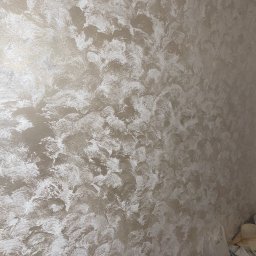 RITA PERETS MegaSklep - Beżowa ściana z dekoracyjną fakturą, imitującą pociągnięcia pędzla. Widoczne nieregularne wzory i połyskujące refleksy światła. W rogu widoczne przedmioty.
