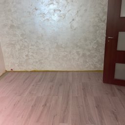 RITA PERETS MegaSklep - Wykończone wnętrze: ściana z dekoracyjną fakturą w odcieniach beżu i brązu, panele podłogowe imitujące drewno oraz fragment brązowych drzwi z przeszkleniami.