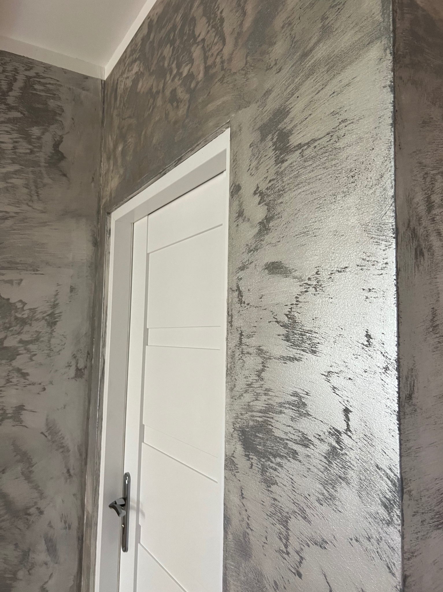 Ściana z efektem dekoracyjnym w odcieniach szarości i srebra, widoczna obok białych drzwi z poziomymi panelami. Tekstura ściany przypomina przecierkę.