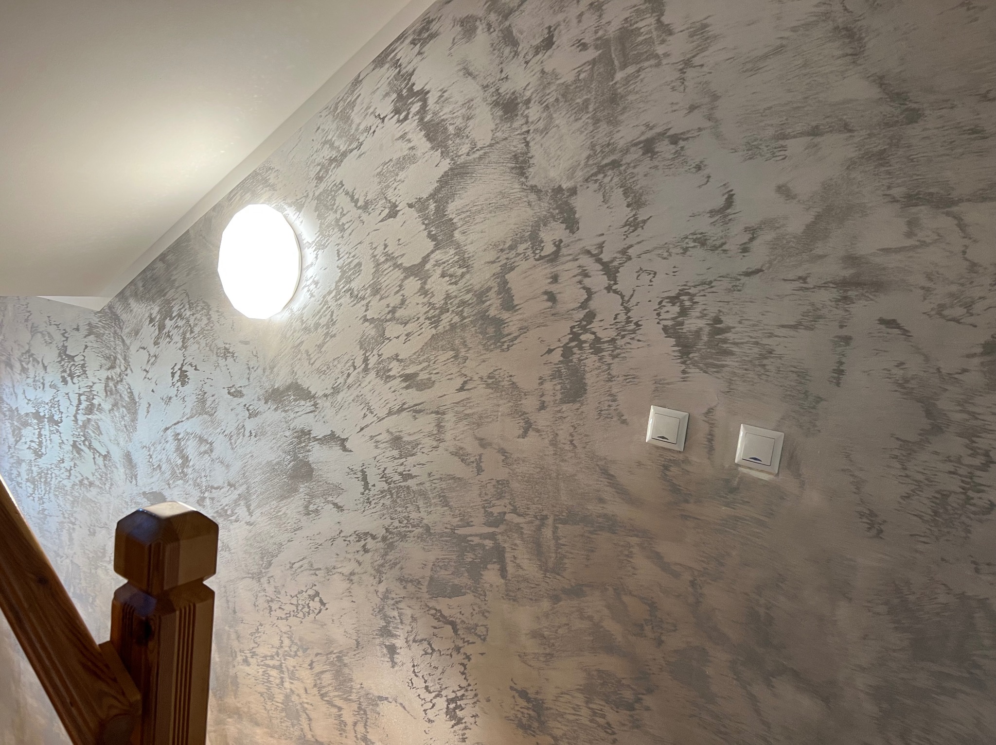 Ściana z efektem dekoracyjnym w odcieniach szarości, oświetlona okrągłą lampą. Widoczne dwa włączniki światła i fragment drewnianej balustrady schodów.