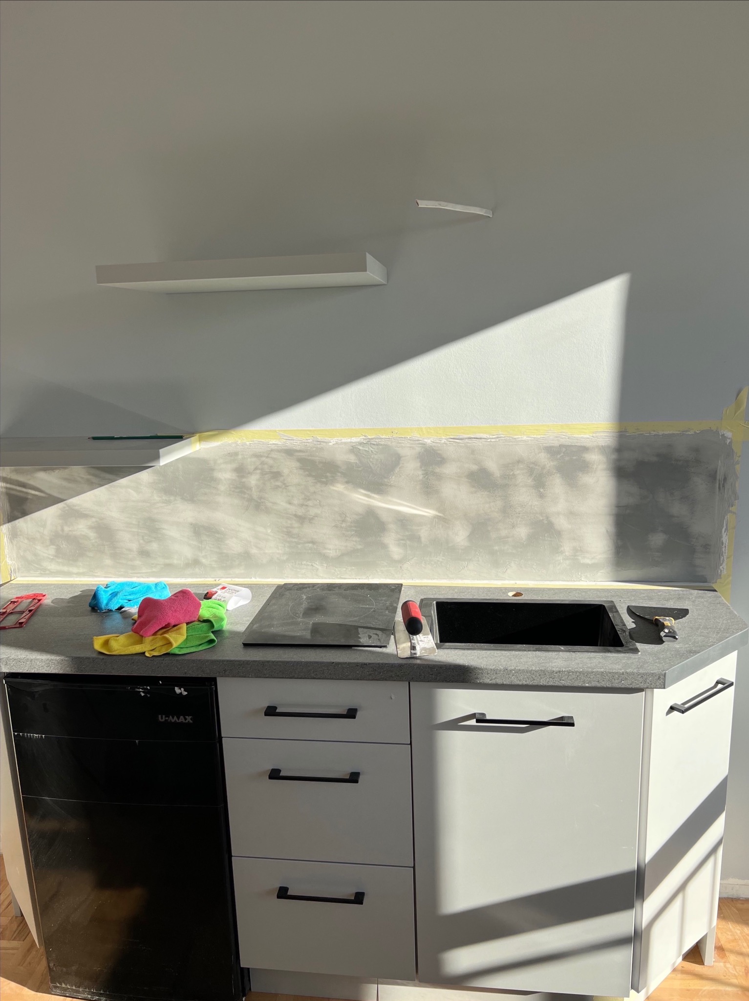 Ściana w kuchni po szpachlowaniu, zabezpieczona taśmą malarską, z widocznymi narzędziami i akcesoriami na blacie. Promienie słoneczne oświetlają ścianę.