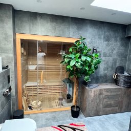 DANIEL DURDA DAN-BUD USŁUGI WYKOŃCZENIOWE - Nowoczesna łazienka z sauną: szare płytki, drewniane wykończenie sauny, roślina doniczkowa. Widoczne akcesoria łazienkowe i fragment toalety. Minimalistyczny design.