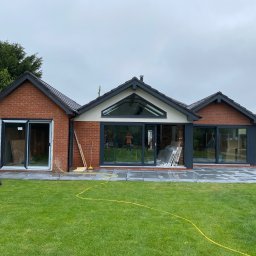 DANIEL DURDA DAN-BUD USŁUGI WYKOŃCZENIOWE - Nowoczesny, parterowy dom z cegły z dużymi oknami i szarymi ramami, otoczony zielonym trawnikiem i nową kostką brukową. Widoczne narzędzia i materiały budowlane.