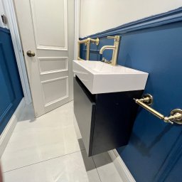 DANIEL DURDA DAN-BUD USŁUGI WYKOŃCZENIOWE - Nowoczesna łazienka: biała umywalka nablatowa z czarną szafką, złota armatura i akcesoria na tle granatowej ściany z dekoracyjnymi listwami.