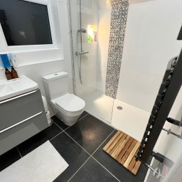 DANIEL DURDA DAN-BUD USŁUGI WYKOŃCZENIOWE - Nowoczesna łazienka po remoncie: szare szafki pod umywalką, biała toaleta, kabina prysznicowa z mozaiką i czarne płytki podłogowe z drewnianym dywanikiem.