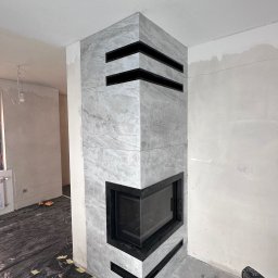 DANIEL DURDA DAN-BUD USŁUGI WYKOŃCZENIOWE - Obudowa kominka z szarych płyt imitujących marmur, z czarnymi wstawkami, w trakcie prac wykończeniowych. Podłoga zabezpieczona folią, widoczne narzędzia.