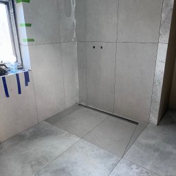 DANIEL DURDA DAN-BUD USŁUGI WYKOŃCZENIOWE - Wykończona łazienka z szarymi płytkami, widoczny odpływ liniowy i przygotowane punkty pod armaturę. Okno zabezpieczone taśmą malarską.