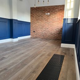 DANIEL DURDA DAN-BUD USŁUGI WYKOŃCZENIOWE - Wnętrze po renowacji: panele ścienne w odcieniach błękitu, ceglana ściana z gniazdkami, drewniana podłoga z kratką wentylacyjną i wiszące lampy. Nowoczesny design.