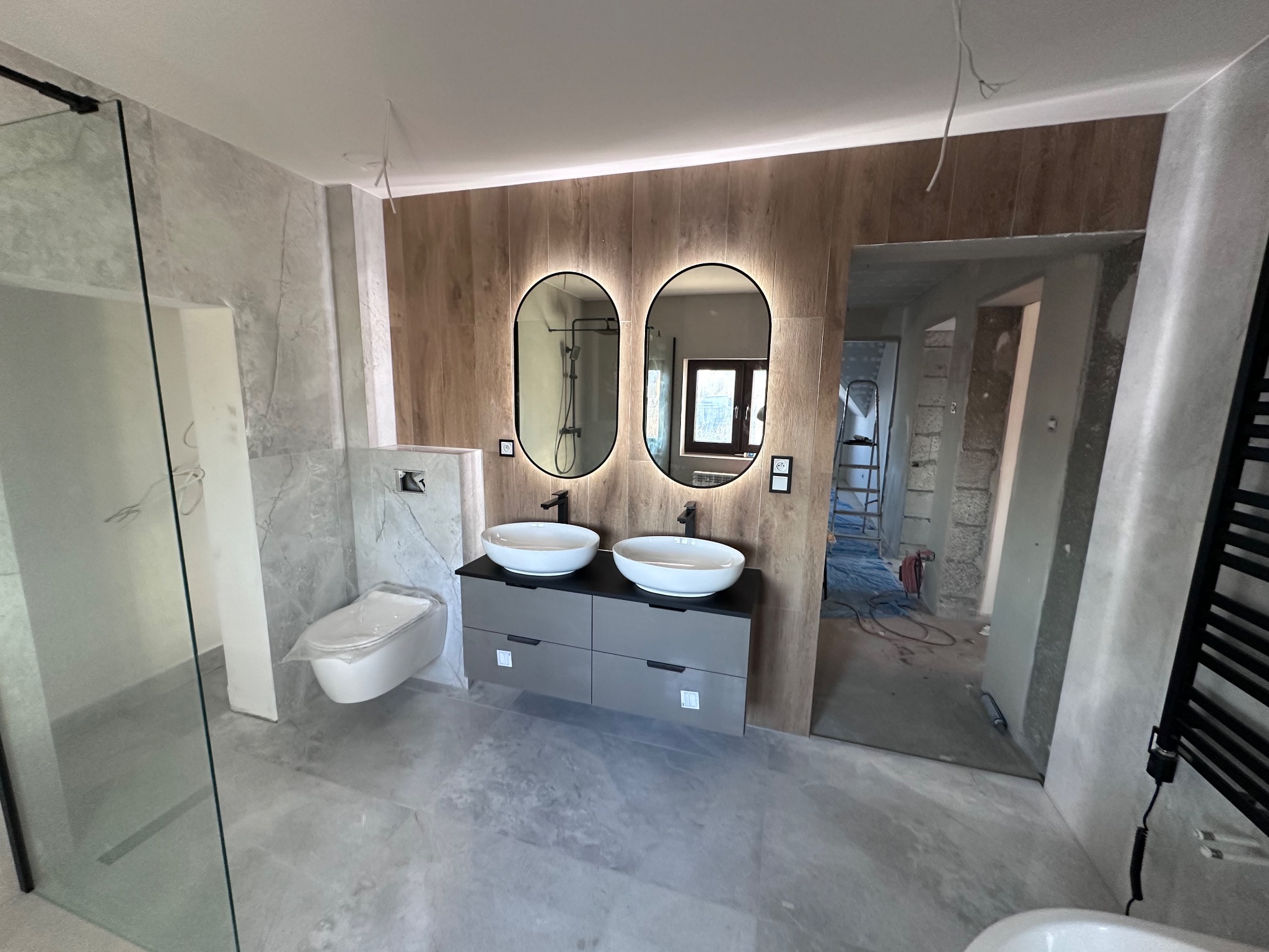 Nowoczesna łazienka w trakcie wykańczania: szare płytki, podwójna umywalka z szafką, owalne lustra z podświetleniem LED, drewnopodobna ściana, widoczne elementy instalacji.