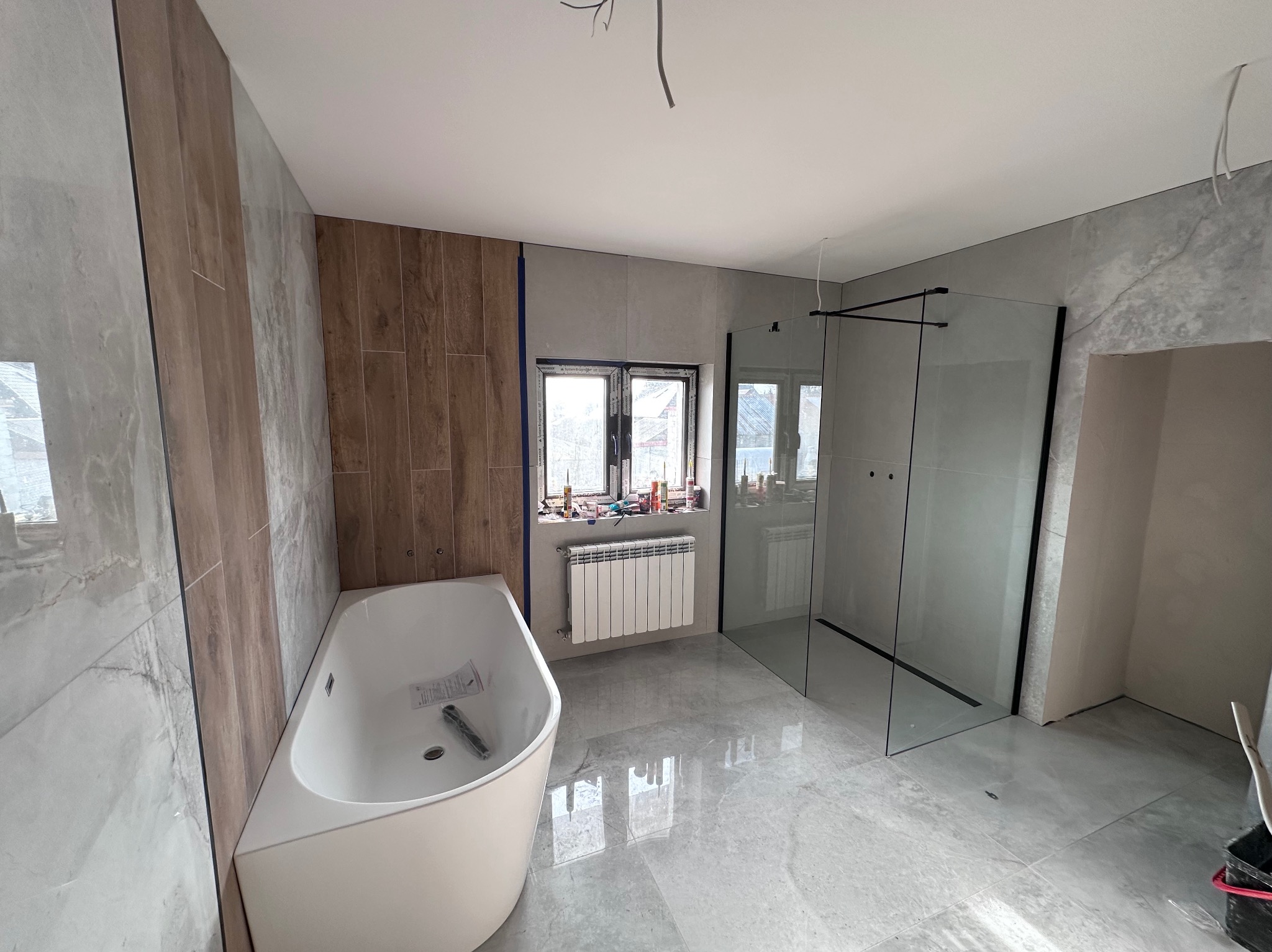 Nowoczesna łazienka po remoncie: szare płytki, drewnopodobne akcenty, biała wanna i kabina prysznicowa typu walk-in z czarnymi profilami. Okno z grzejnikiem.