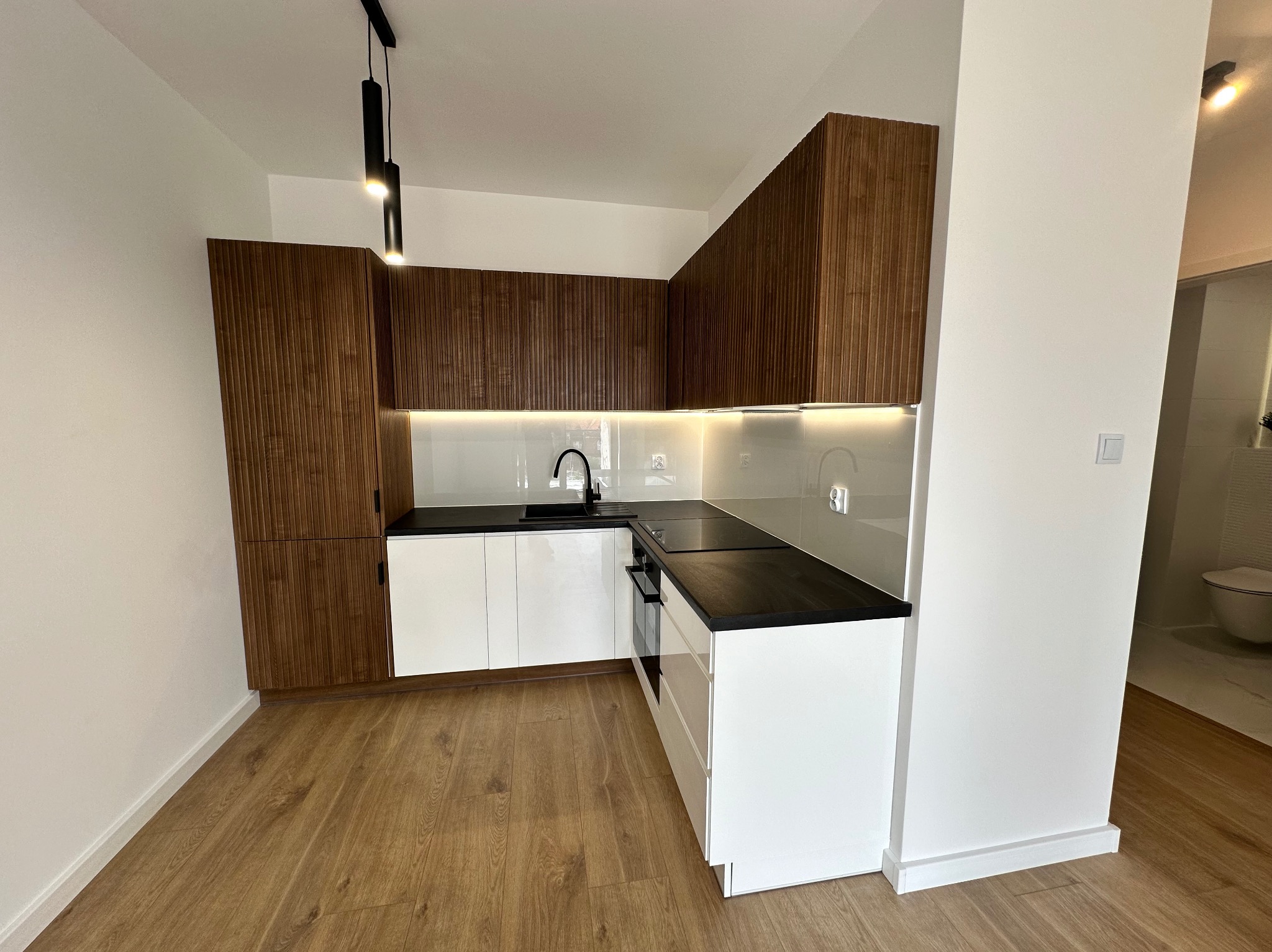 Nowoczesna kuchnia z drewnianymi szafkami górnymi i białymi dolnymi, czarnym blatem, oświetleniem LED i drewnianą podłogą. Widok na łazienkę w tle.