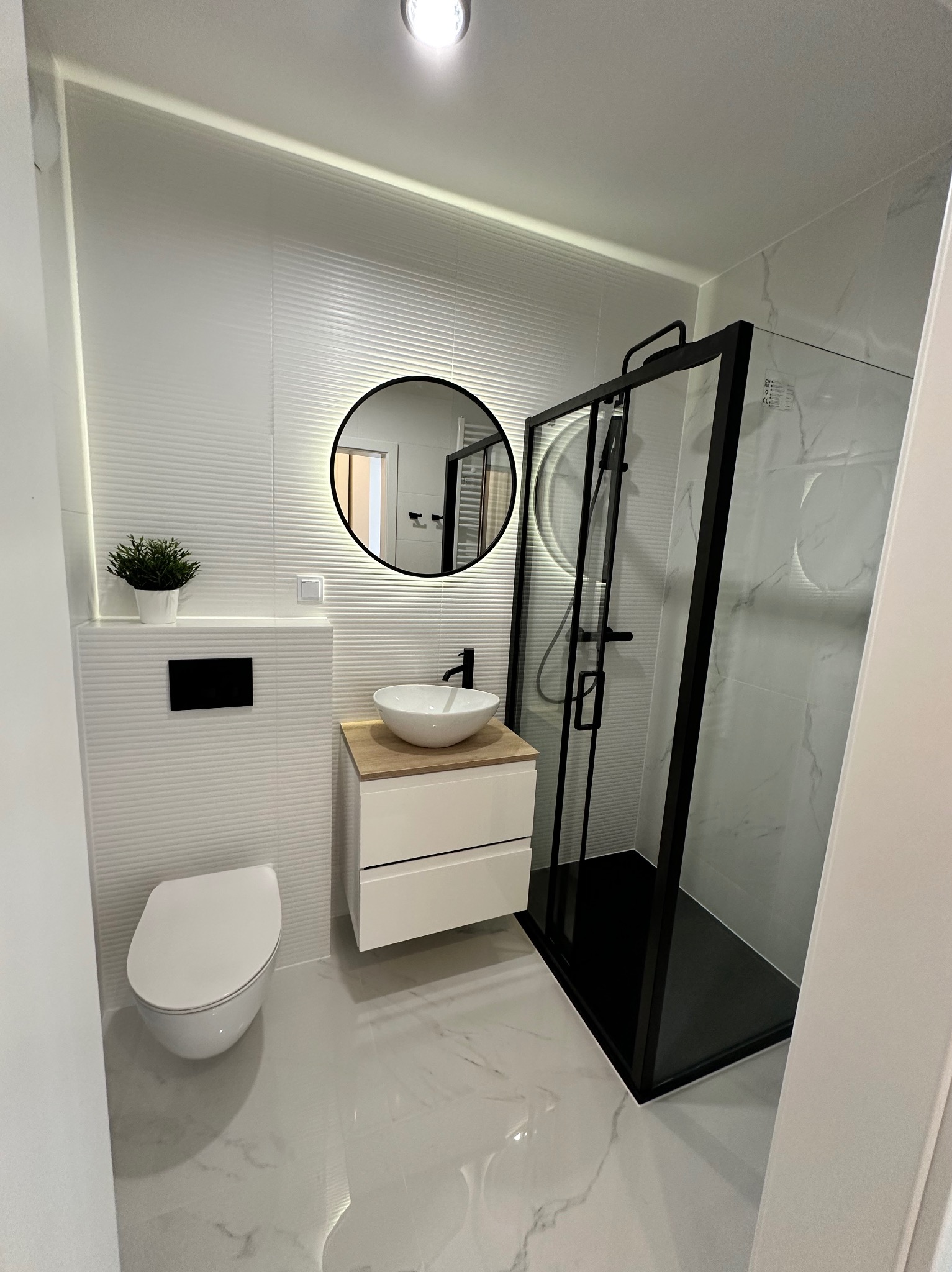 Nowoczesna łazienka w bieli: kabina prysznicowa z czarną ramą, okrągłe lustro z podświetleniem, umywalka nablatowa i podwieszana toaleta. Minimalistyczny design.