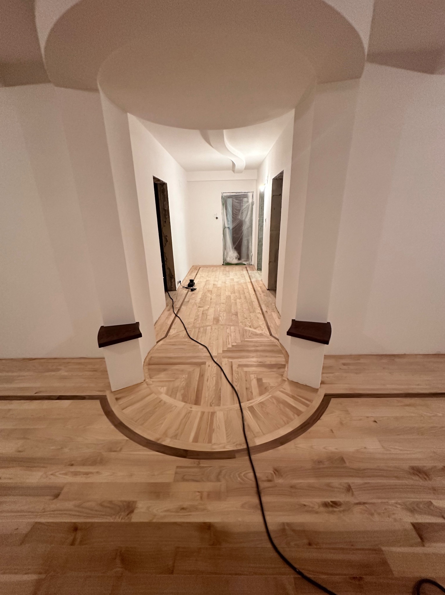 Parkiet ułożony w korytarzu z dekoracyjnym wzorem, widoczne elementy wykończeniowe ścian i sufitu. Kabel elektryczny na podłodze sugeruje trwające prace.