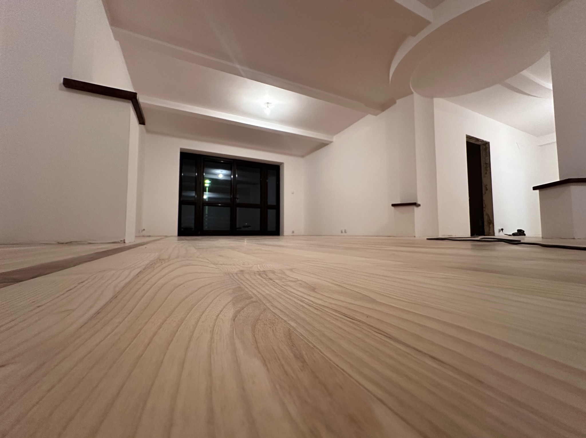 Jasna, drewniana podłoga po renowacji w przestronnym wnętrzu z białymi ścianami i czarnymi ramami okiennymi. Widok z niskiej perspektywy podkreśla strukturę drewna.