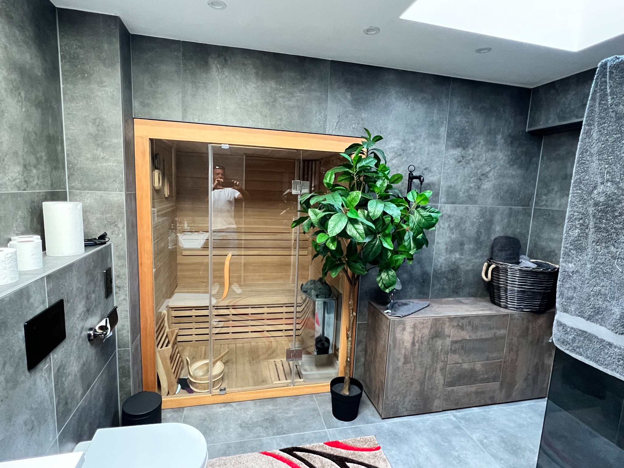 Nowoczesna łazienka z sauną: szare płytki, drewniane wykończenie sauny, roślina doniczkowa. Widoczne akcesoria łazienkowe i fragment toalety. Minimalistyczny design.