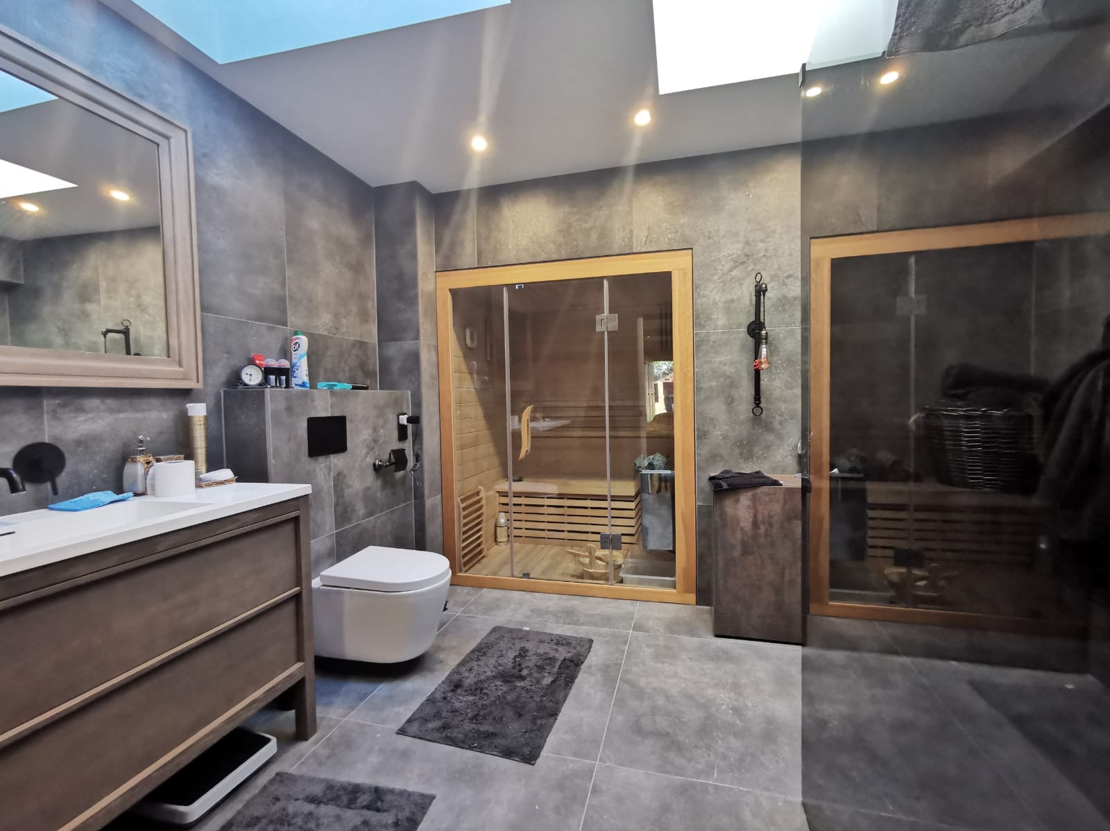 Nowoczesna łazienka z sauną: szare płytki, drewniane akcenty, oświetlenie punktowe i duże lustro nad umywalką. Minimalistyczny design z dbałością o detale.