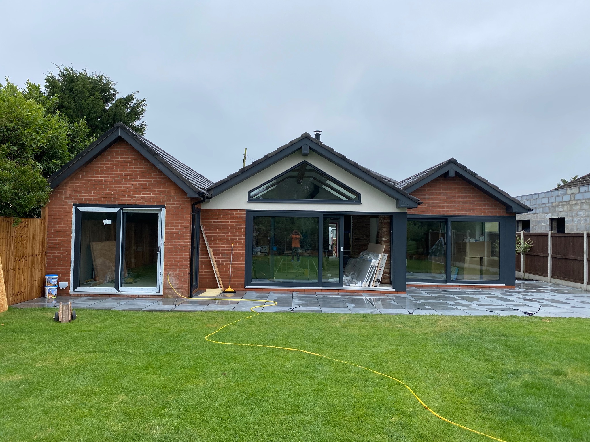 Nowoczesny, parterowy dom z cegły z dużymi oknami i szarymi ramami, otoczony zielonym trawnikiem i nową kostką brukową. Widoczne narzędzia i materiały budowlane.